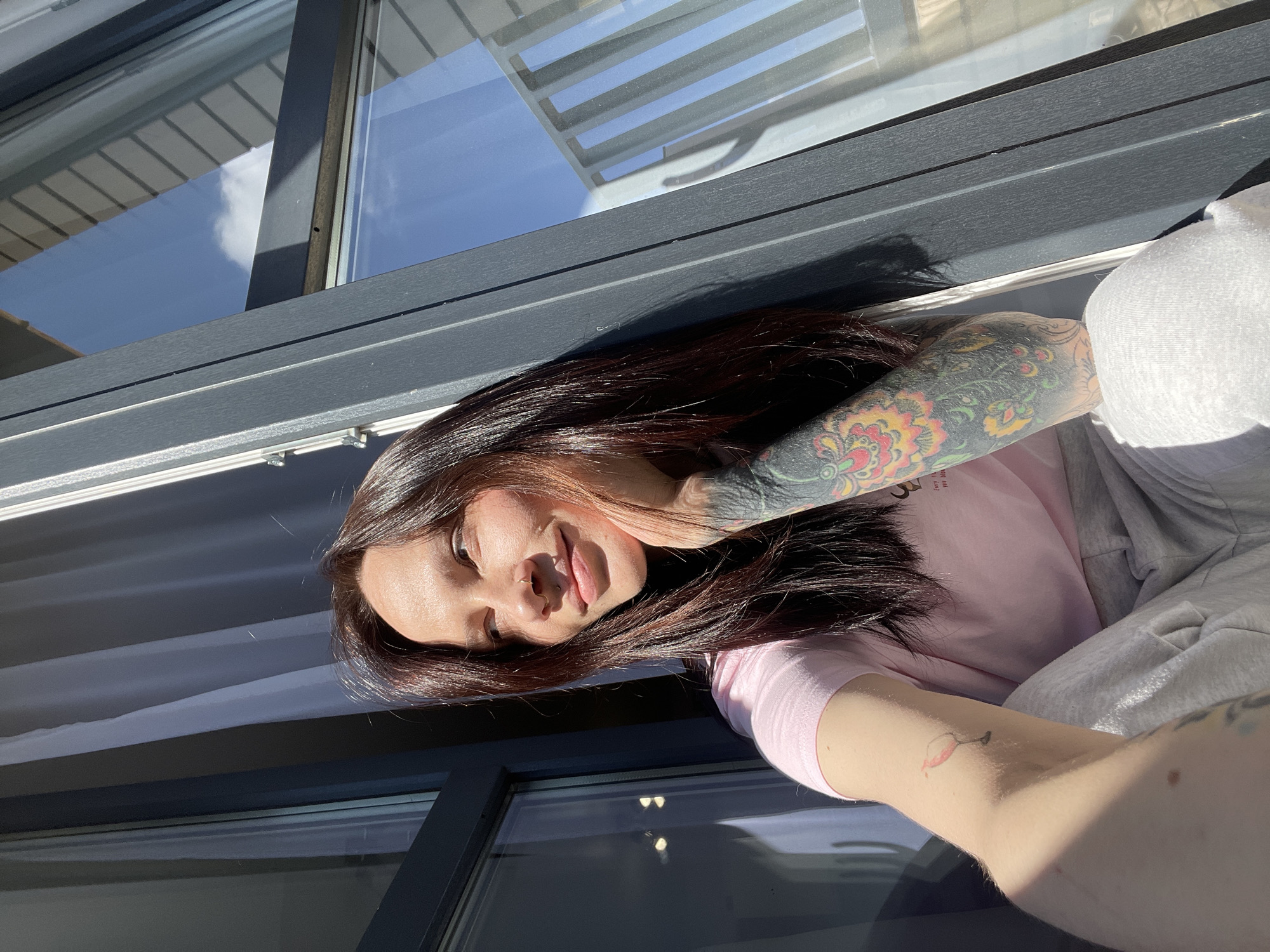 A young woman with dark brown hair is pictured in an indoor setting with sunlight streaming through a window. She is wearing a light pink t-shirt and a grey sweatshirt. The woman has visible tattoos on her arms. The background shows a window with a dark frame and visible reflections of the sky and clouds. The location is unknown, there is nothing in the image that would indicate it is in Izhevsk, Russia.