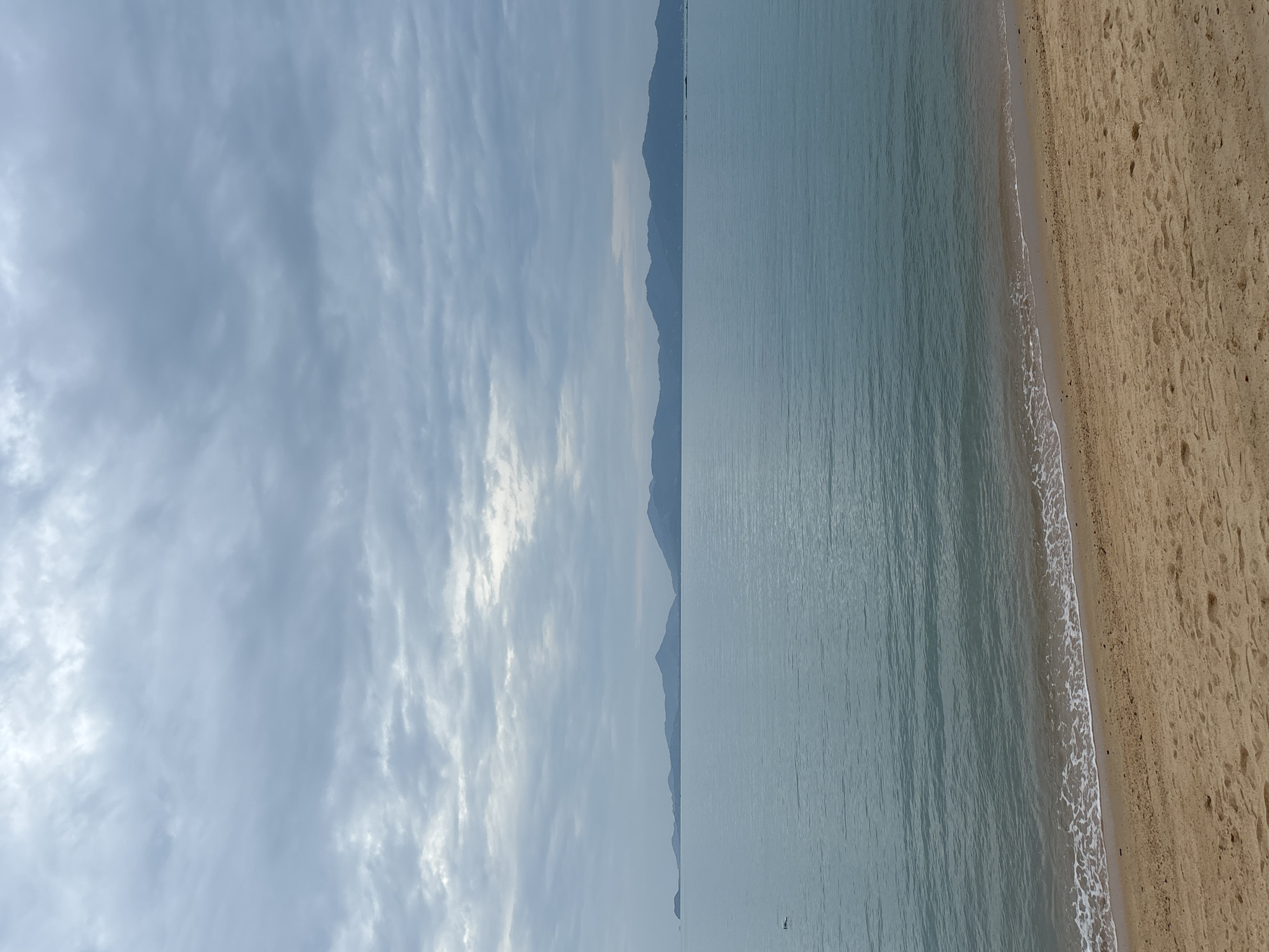 The image depicts a beach scene in Baan Bo Phut, Thailand. The sky is overcast with cloud formations. A distant mountain range is visible across the water. Calm, grey-blue water transitions into a sandy beach in the foreground. Small waves are visible where the water meets the shoreline. The beach is beige with footprints in the sand. There appear to be no people visible in the image. There is also a small boat in the water, near the far shore.