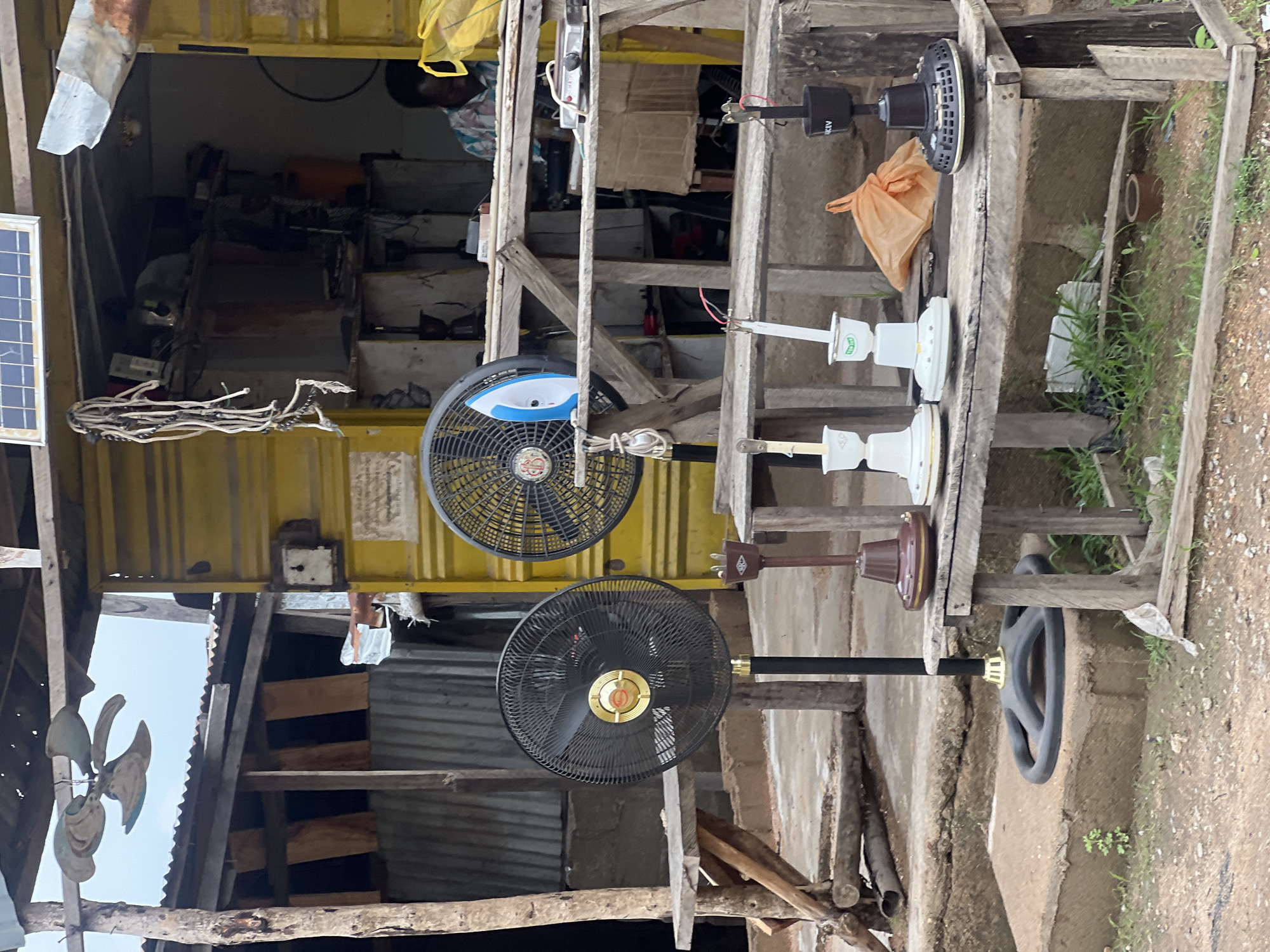 The image depicts an outdoor repair shop in Jalingo, Nigeria, specializing in electrical appliances, particularly fans. Several fans are visible, some in working condition and others in various states of repair. In the foreground, two large black standing fans are displayed. To their right, on a wooden shelf, are several smaller ceiling fan components and a single iron. A man is seated inside the shop, appearing to be working on an appliance. The shop itself is a small, open-air structure with a yellow corrugated metal facade. The surrounding environment is dusty and appears to be a roadside market or workshop. The overall impression is one of a functional, albeit somewhat cluttered, local business catering to everyday electrical needs.