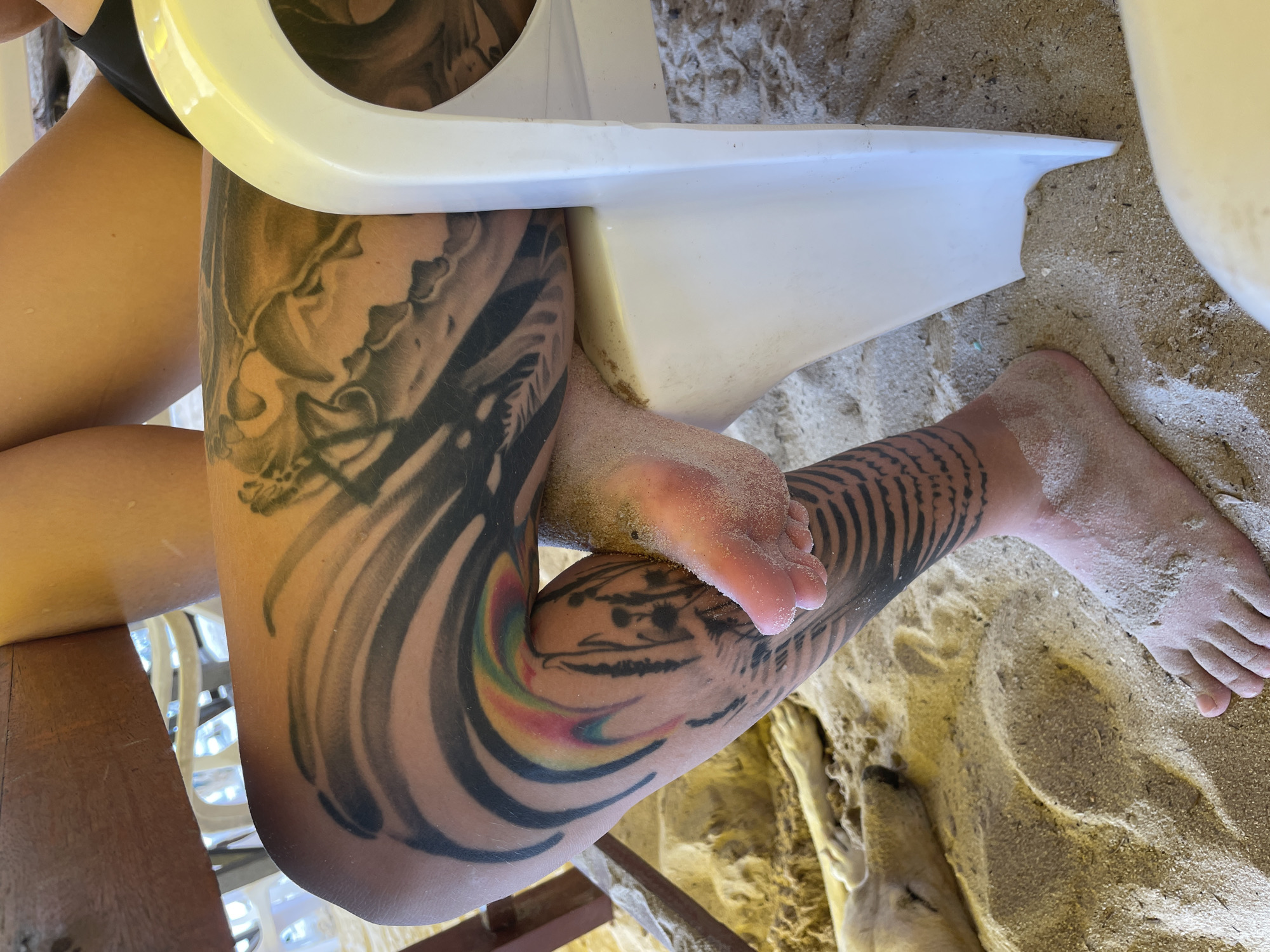 The image shows a person sitting on a white plastic chair on a sandy beach in Choeng Thale, Thailand. The person has a tattoo on their right thigh and another tattoo on their lower right leg. Both feet are visible and partially covered in sand. The beach sand is beige and contains some small pebbles. A small portion of a wooden structure is visible beneath the chair. The weather appears to be sunny.