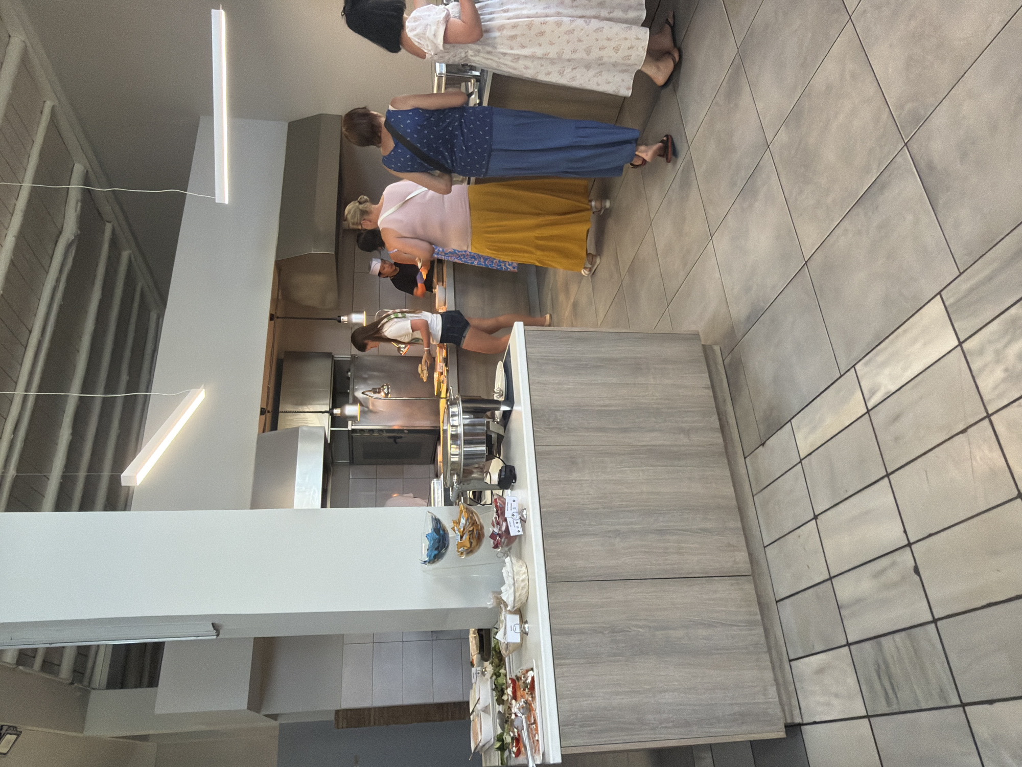In Georgioupoli, Greece, a buffet line is depicted in a bright, modern dining area. Several individuals are observed selecting food from a counter filled with various dishes and serving containers. A large buffet warmer is prominently positioned on the counter, alongside bowls of snacks and wrapped items. The floor is tiled in a light gray, and the walls are a neutral tone. The overall scene suggests a casual dining setting, possibly a hotel or restaurant.