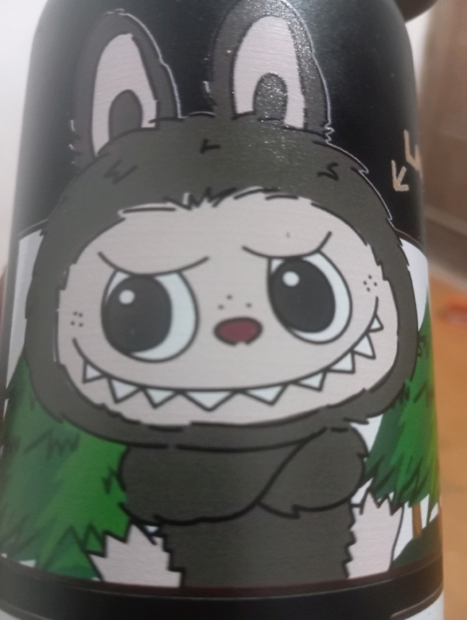 The image shows a close-up of a black cylindrical object, possibly a water bottle, decorated with a cartoon illustration. The illustration depicts a creature with large, round eyes, a wide mouth with sharp teeth, and bunny-like ears. The creature is wearing a hooded outfit with paws. Green, stylized trees are visible on the sides of the object. A small, light-colored symbol resembling an arrow or a checkmark is present on the upper right side of the object. The background is blurred and appears to be an indoor setting. There is no visible indication of Kutasari, Indonesia, in the image or its context.