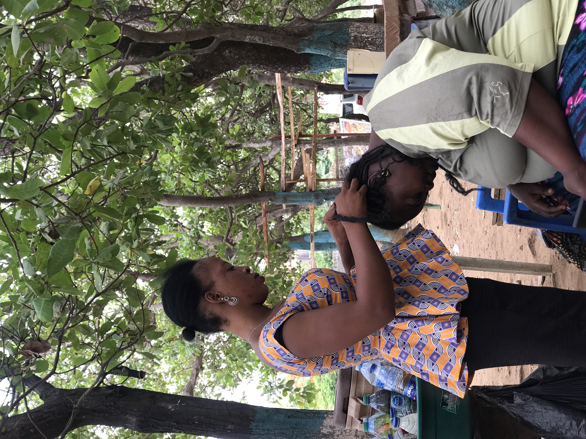 In an outdoor setting in Abuja, Nigeria, a woman in a patterned orange and black top stands facing left, braiding the hair of another woman who is bent over. The braider's earrings and a necklace are visible. A table in front of her holds several water bottles. The other woman is wearing a striped green and gray shirt and is seated on a blue plastic chair. Trees surround the area. A wooden structure is visible in the background. The ground is dirt.