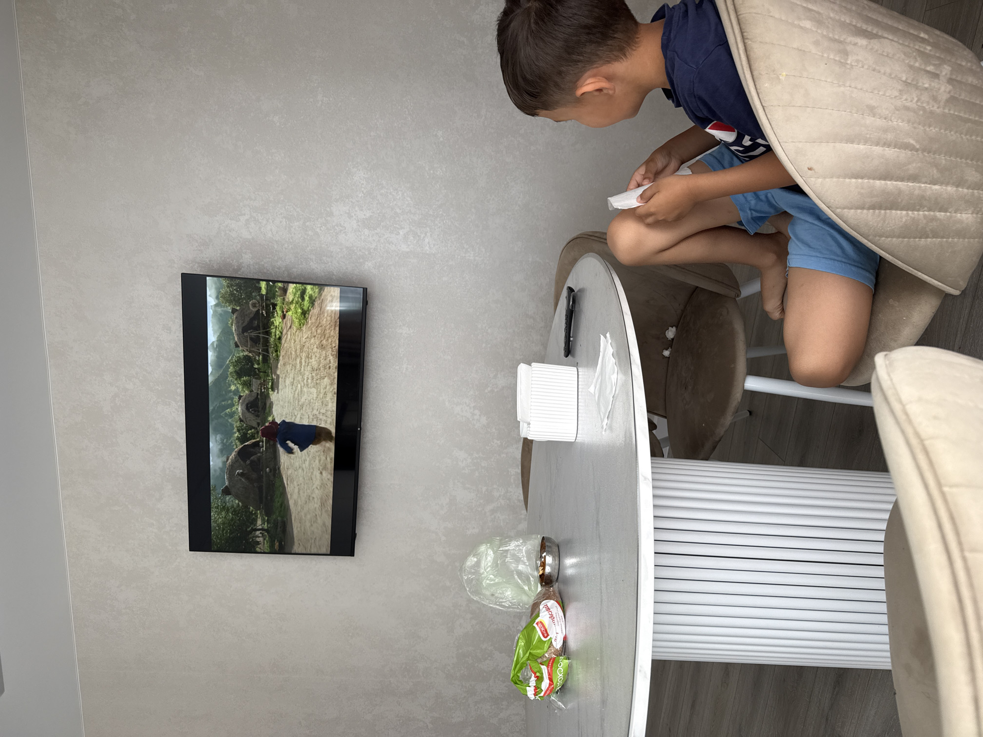 The image shows a young boy sitting at a round table in a room. The boy is wearing a blue t-shirt and blue shorts, and he is holding a white object, possibly a remote control, in his hands. He is looking down at the object with a focused expression.
The table is round and has a white base with vertical slats. On the table, there are several objects, including a white tissue dispenser, a plastic bag, and a green and red package with some food items inside. A TV screen is mounted on the wall to the left of the boy. The TV displays an image of what appears to be a scene from a movie or show, featuring animals and a person.
The room has light-colored walls with a textured finish. The floor is made of wooden planks. The boy is sitting on a chair that is partially covered by a beige upholstered piece. The overall setting suggests a casual indoor environment, possibly a dining area or living room. The lighting in the image is bright, indicating it might be daytime. There are no clear indicators of the specific time of day or weather conditions. The scene is static and appears to capture a moment of quiet activity.