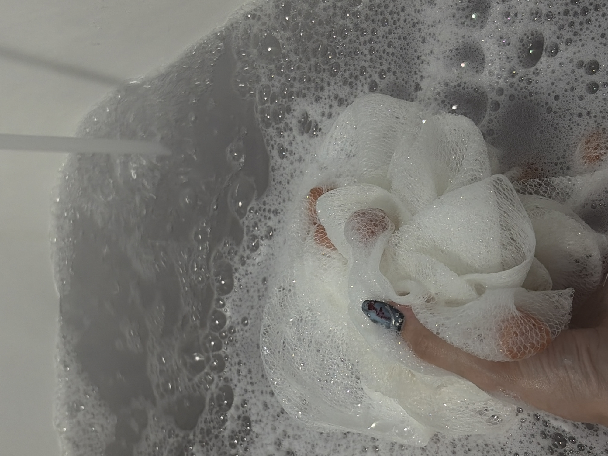The image depicts a close-up view inside a bathtub filled with foamy water. A hand holds a white loofah sponge amidst the bubbles. Water is flowing from a tap on the left side of the frame. The environment appears to be a bathroom.