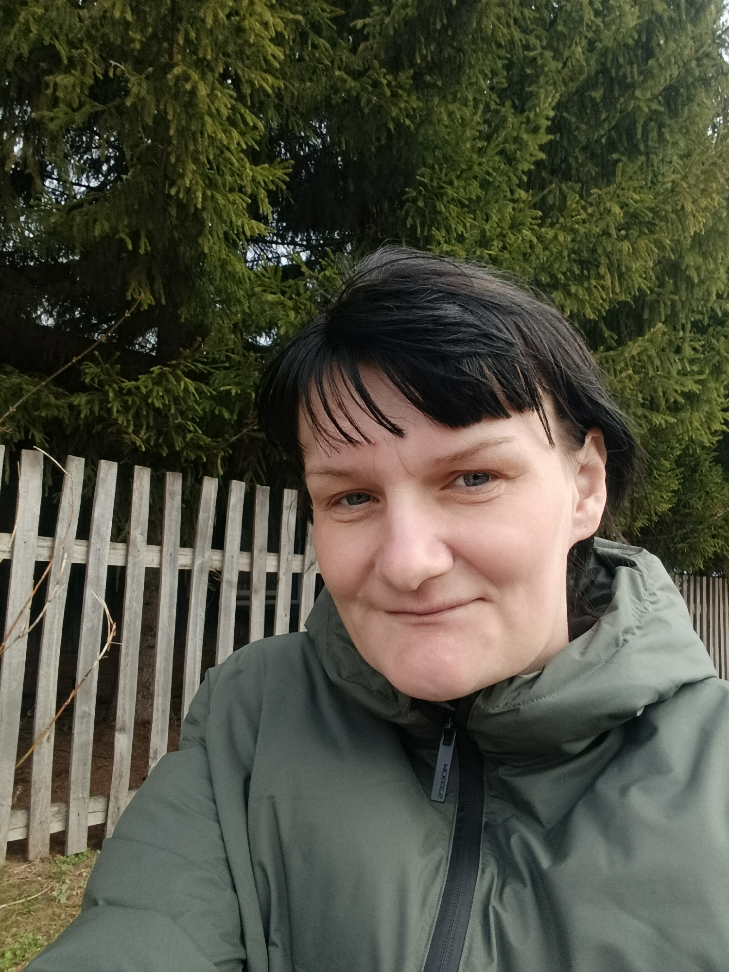 A woman stands in front of a wooden fence and evergreen trees. She is wearing a green hooded jacket and has short, dark hair with bangs. The location is in an unidentified area of the city of Unknown, Unknown. The weather appears to be overcast or cloudy.