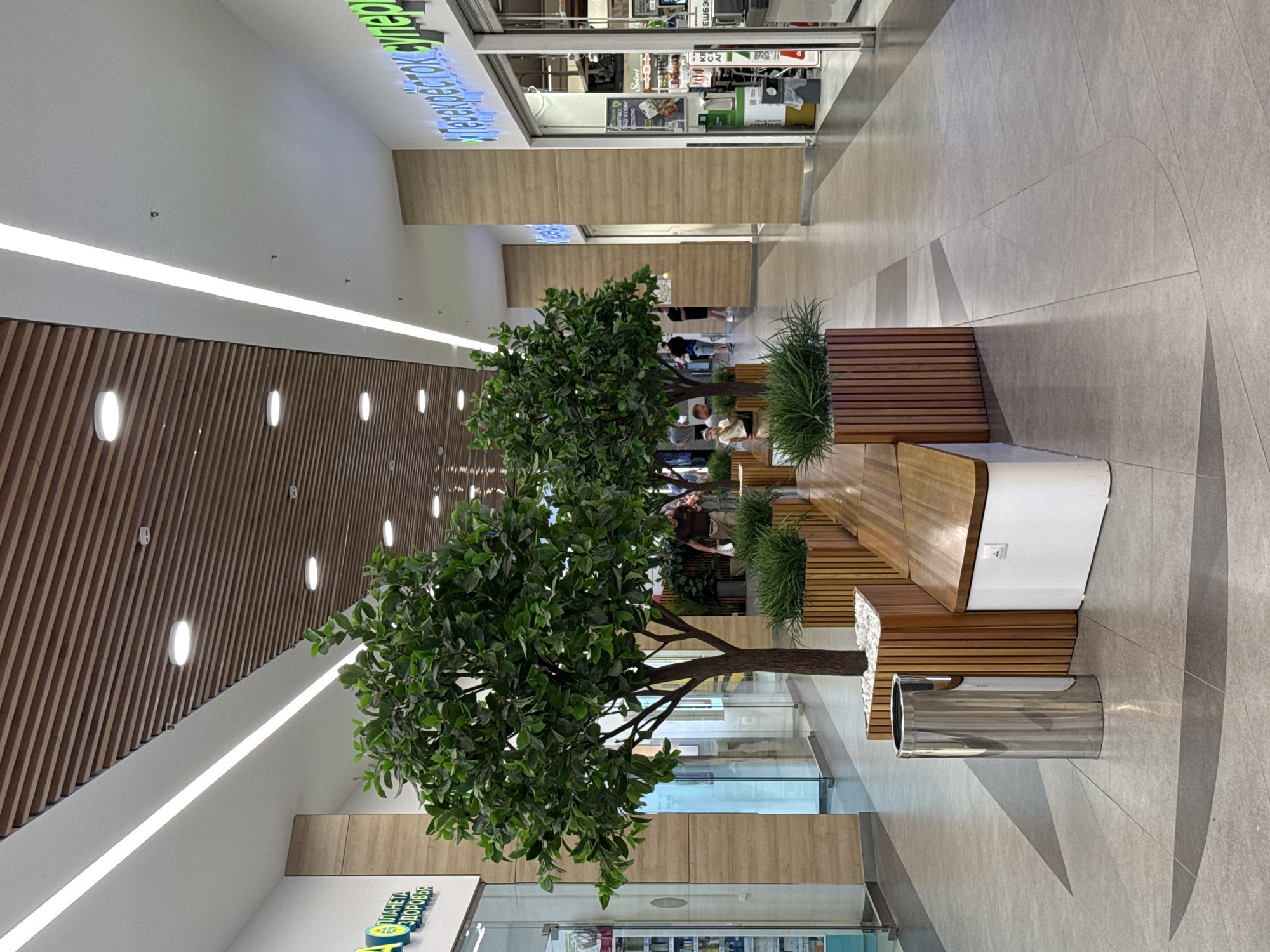 The image shows an indoor shopping mall corridor with artificial trees and benches. The architecture features wooden slat ceilings and light-colored walls. In the foreground, there is a cylindrical stainless steel trash can. The corridor is lined with stores, one of which has a prominent blue neon sign that reads "Перекрёсток Супермаркет" (Perekrestok Supermarket). There are several people walking and standing in the background. The floor is tiled with a grey patterned design. The overall impression is a clean, modern, and well-lit commercial space. This specific location is identified as being in Izhevsk, Russia, based on the recognizable "Perekrestok" supermarket chain signage.