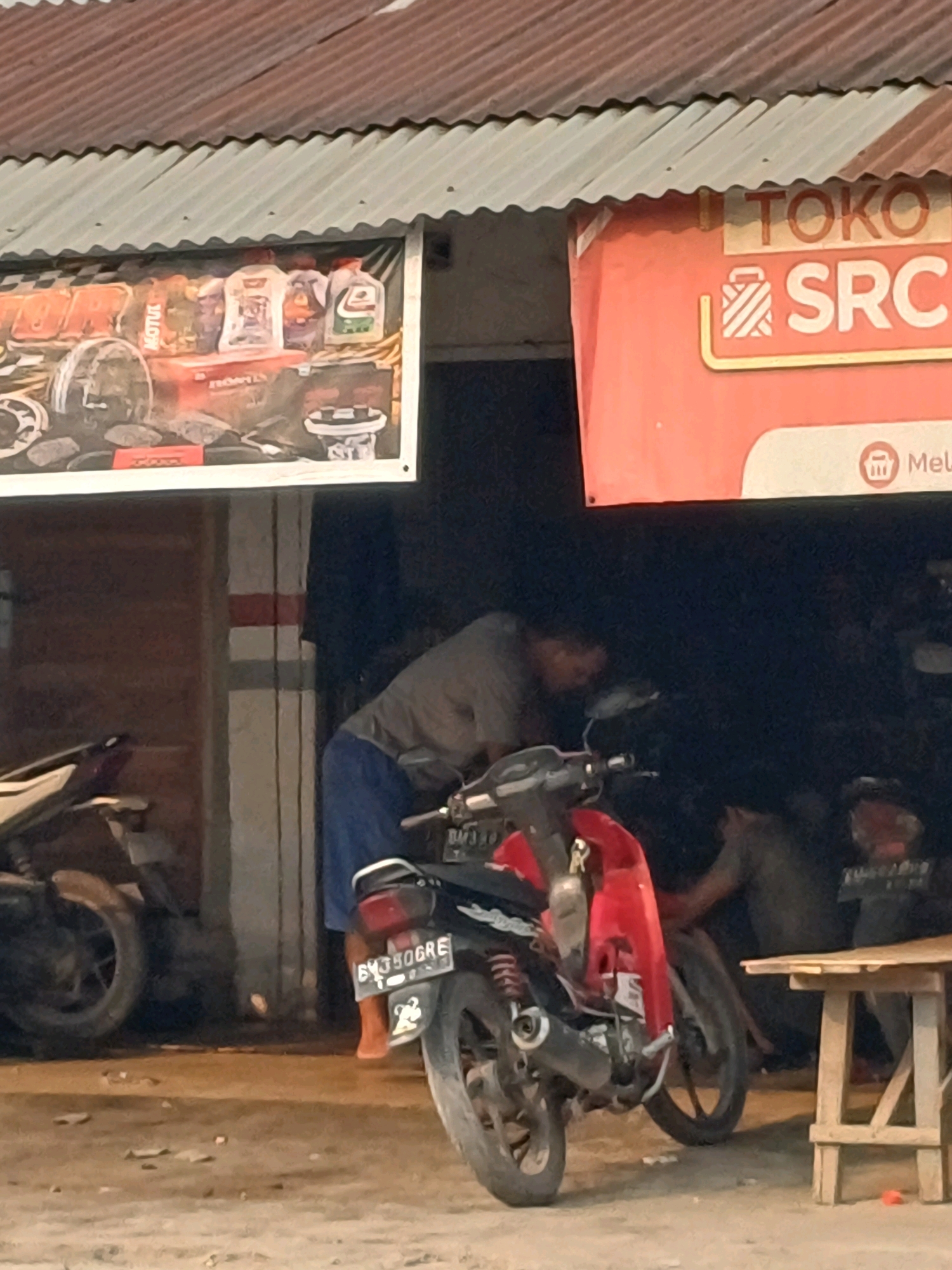This is a photograph taken outdoors, likely in Dumai City, Indonesia, depicting a street-level scene in front of a shop. The shop has a corrugated metal roof extending over a banner that advertises "TOKO SRC." Below the banner, the shopfront is open, revealing the interior where a person in a grey t-shirt and blue shorts is working on a red and black motorcycle. Another person is partially visible in the background. Two motorcycles are parked in front of the shop, one in the foreground partially obscuring the other. A wooden table is also present. The ground is paved with uneven concrete, and debris is scattered on the surface. The lighting suggests it is either late afternoon or early evening, casting shadows and creating a dimly lit interior. The overall impression is of a busy, everyday scene at a local motorcycle repair shop or parts store.