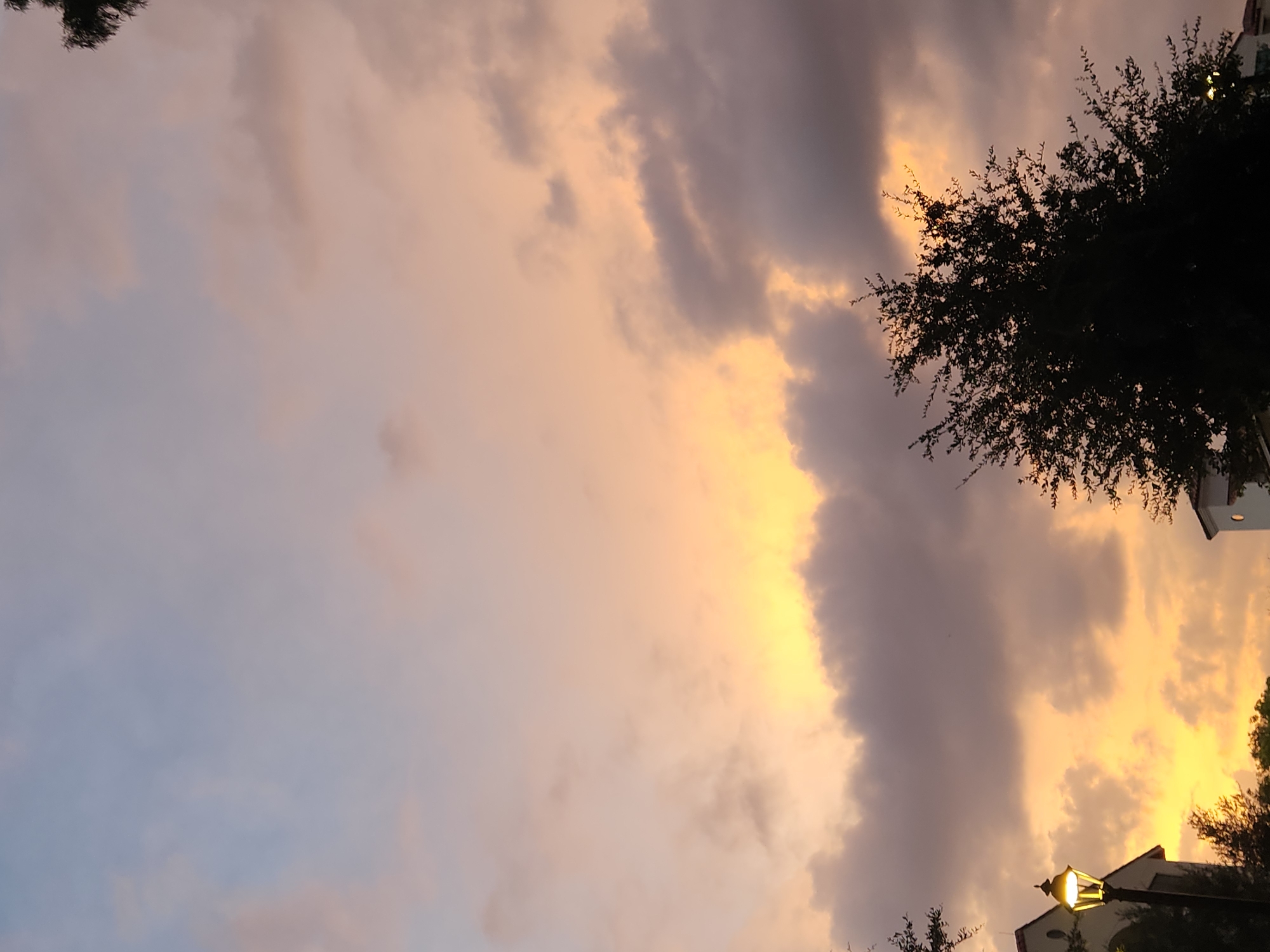 A nighttime exterior shot captures a sunset with hues of orange, pink, and purple filling the sky, with clouds dispersed throughout. In the lower left quadrant, a lit lamppost stands beside a building with architectural details suggestive of a courtyard or residential complex. In the lower right quadrant, another building is partially visible. Silhouetted trees frame the scene from the bottom edges. The image appears to have been taken in Winter Park, United States of America.