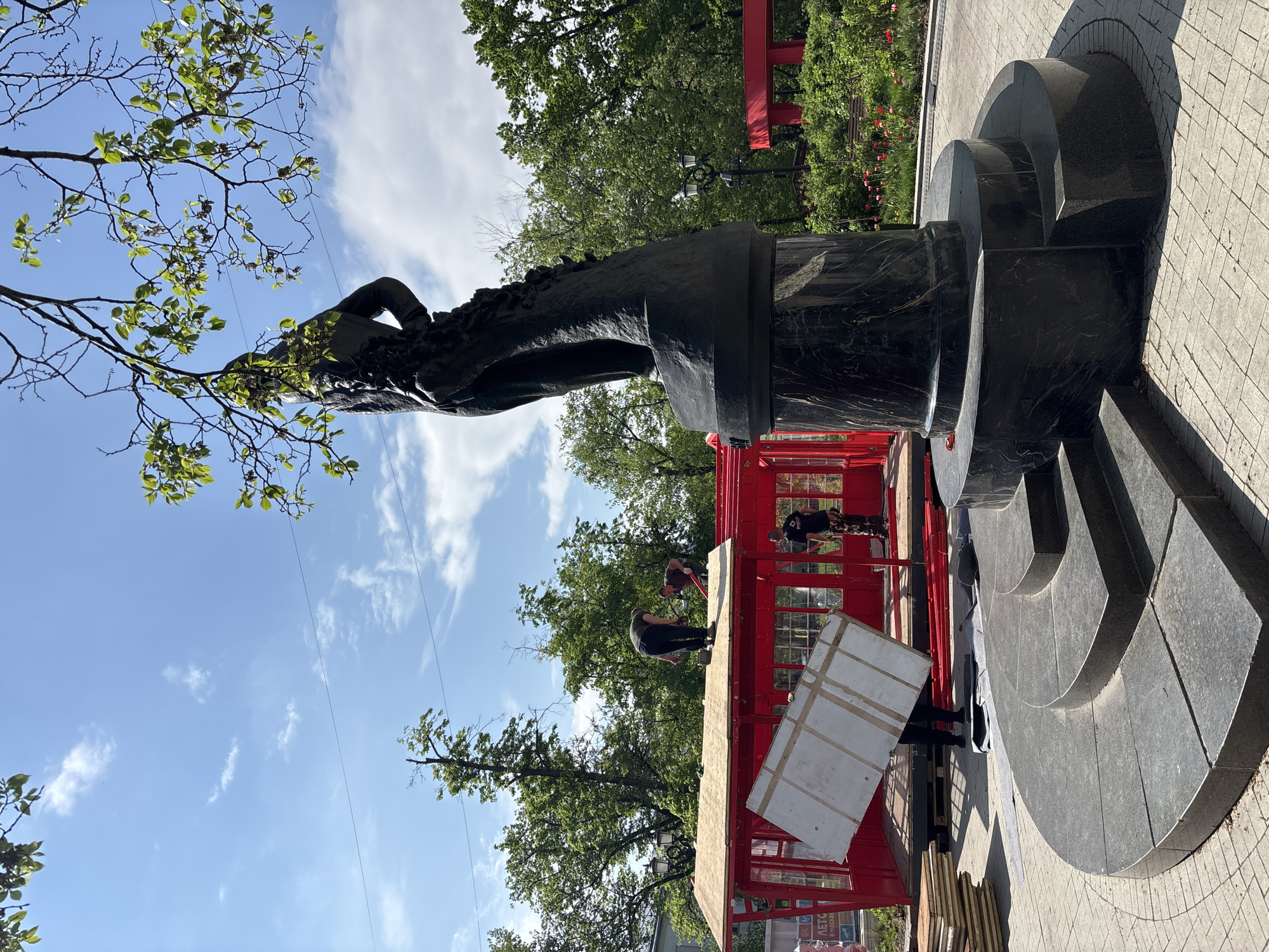 The image shows a statue in Moscow, Russia, being worked on. The statue is made of a dark material, likely bronze or a similar metal. It sits atop a circular, tiered base constructed from dark gray stone. Two people are standing on a red platform, which appears to be some kind of scaffolding or structure built around the statue. One person seems to be working on the statue itself, while the other holds up a large, rectangular object, possibly a panel or piece of equipment. The sky is blue with some scattered clouds, and there are trees in the background. The statue is positioned near a paved area.