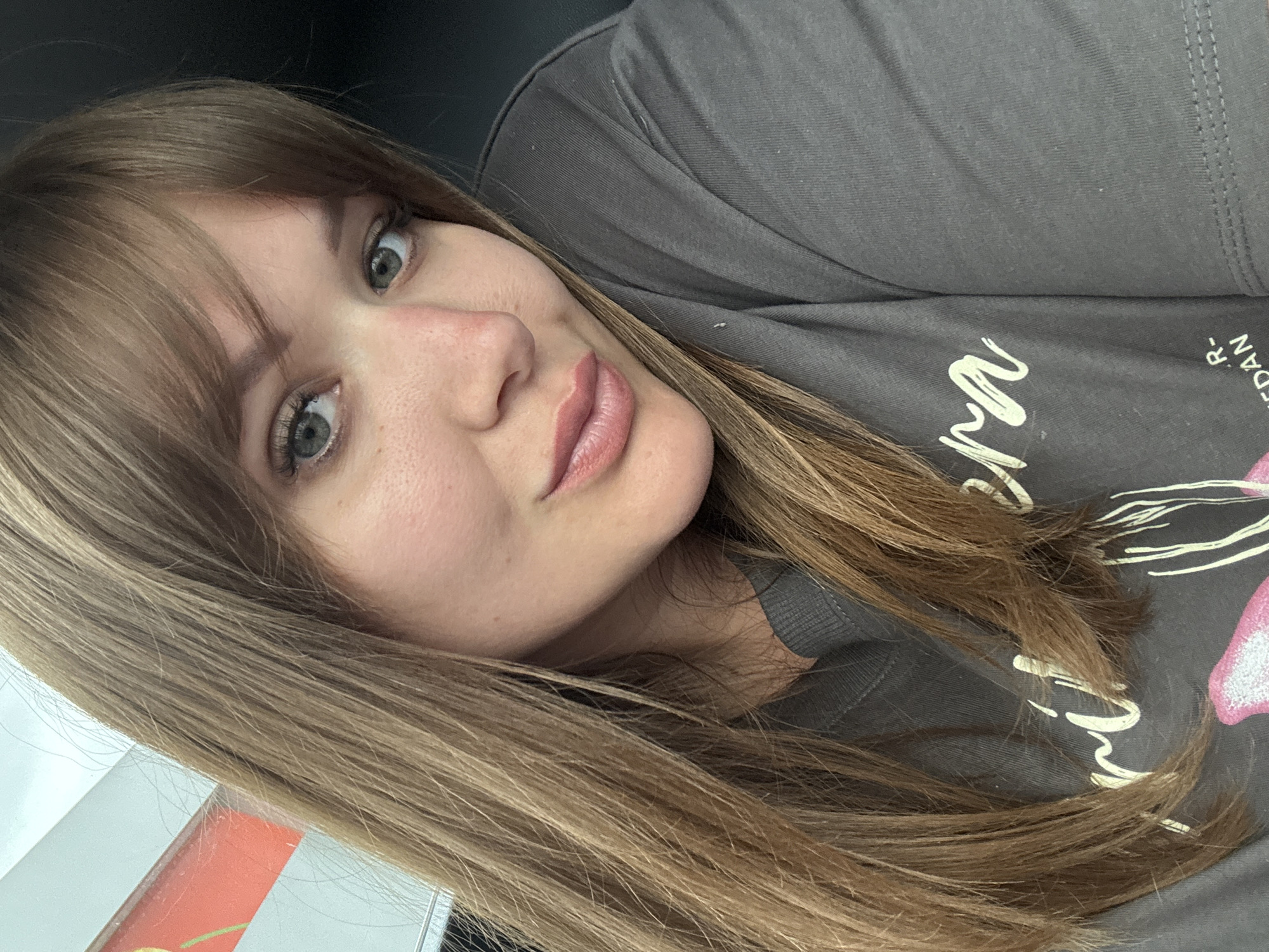 The image is a close-up selfie of a woman. She has fair skin, light brown hair with bangs, and light-colored eyes. She is wearing a gray t-shirt with a graphic design on it. The background is out of focus but appears to be a dark or shadowed area. There is a hint of a window and an object, possibly packaging, with an orange and white design on the lower left of the image. No information about the city of Steblitskii, Russia is present in the image.