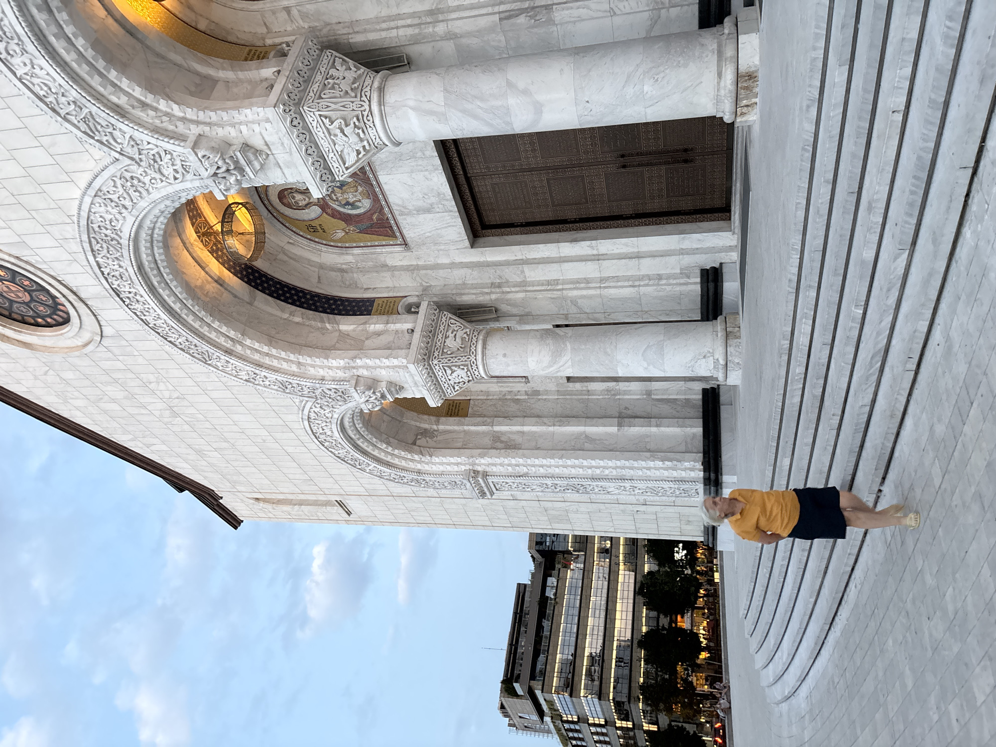 The image shows the exterior of the Church of Saint Sava in Belgrade, Serbia. A woman with white hair, wearing an orange polo shirt and dark shorts, walks down a wide, curved set of marble stairs leading to the church's entrance. The church facade is made of white marble with ornate arches, columns, and a large bronze door. Above the door, a mosaic depicts a religious figure. In the background, a modern glass-and-steel building is visible, reflecting the sky. The sky is a pale blue with scattered clouds. The overall scene appears to be set during twilight or early evening.