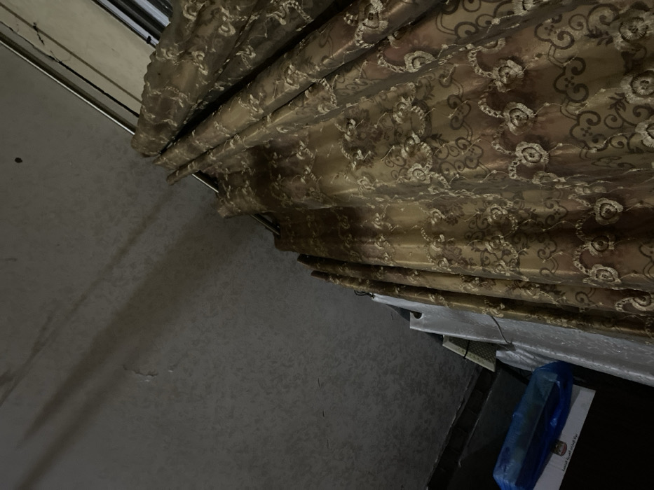 A section of heavy, patterned drapes in brown and gold tones cascades from a window or doorway. The fabric is ornately decorated with a swirling floral design. Beneath the drapes, a dark, cluttered area reveals a table with items partially covered in plastic. The adjacent wall is pale gray, showing some staining or uneven texture. The perspective is from a low angle, emphasizing the drapes' fullness and height.
