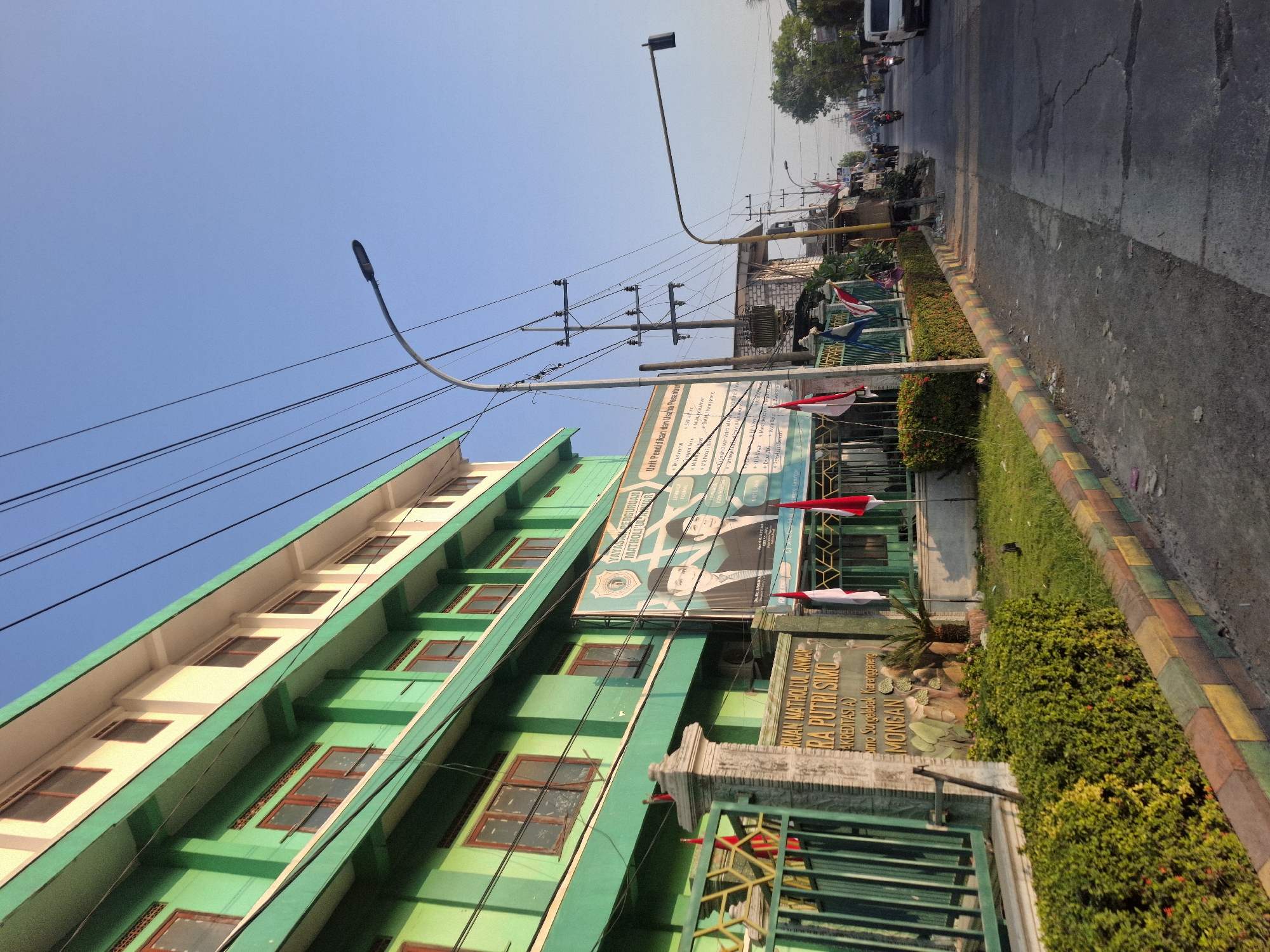 The image captures a street scene in Sungelebak, Indonesia, under a clear blue sky. On the left, a prominent green and cream-colored building with multiple floors and windows lines the street. A metal fence with decorative elements and a stone pillar stands in front of the building. A large banner featuring two men in formal attire is displayed on the building, along with Indonesian flags. Adjacent to the building is a well-maintained strip of green lawn and bushes, bordered by a colorful concrete curb. The street itself is paved and shows signs of wear, with cracks and patches. Several utility poles and streetlights line the side of the road. In the distance, the street continues, with more buildings and trees visible. Vehicles, including motorcycles and a white van, are present on the street, suggesting ongoing traffic. The overall impression is of a well-established urban area bathed in natural light, likely during the daytime.