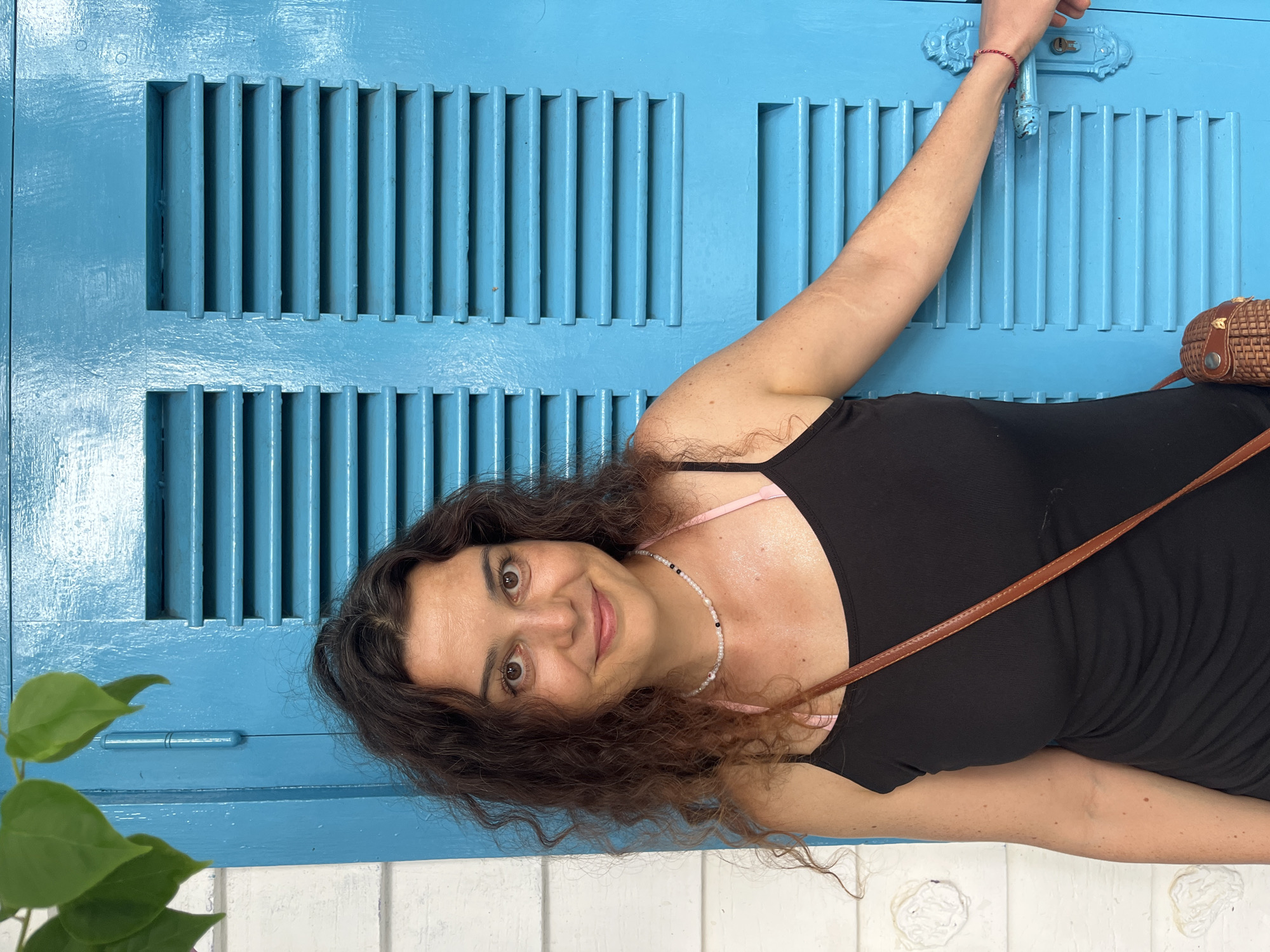 A woman with dark, curly hair and brown eyes is standing in front of a bright blue louvered door in Phu Thuy Ward, Vietnam. She is wearing a black tank top, a beaded necklace, and a small woven bag is slung across her body. Her right arm is extended, and her hand is reaching for the door handle. Green leaves are visible in the upper left corner of the image, suggesting an outdoor setting. The wall to the left of the door is white with decorative elements.