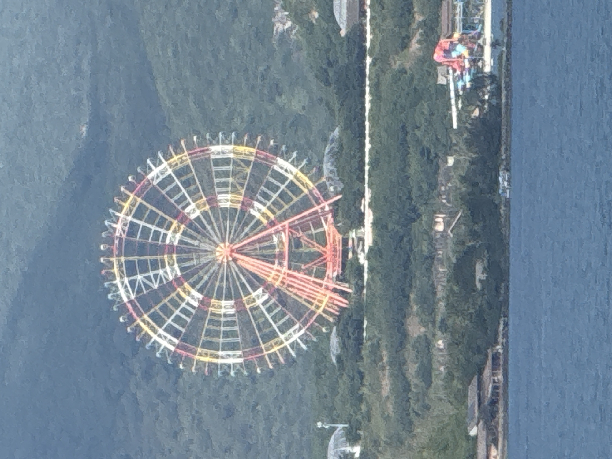 The image shows a large Ferris wheel in the city of Nha Trang, Vietnam. The Ferris wheel is colorful, with red, yellow, and white accents. It is located on a hill overlooking the city and the ocean. In the foreground, there is a body of water, likely the ocean, and some buildings and trees on the shore. There are no people visible in the image. The Ferris wheel appears to be part of an amusement park or resort. The overall scene is a scenic view of a coastal city with a prominent amusement park attraction.