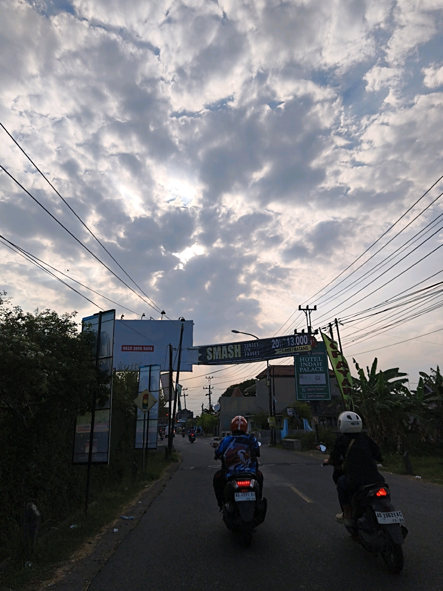 The image captures a street scene in Desa Ngesrep, Indonesia, under a dramatic, cloud-filled sky. Two individuals are riding motorcycles in the foreground, traveling away from the viewer down a paved road. The motorcycle on the left has a rider wearing a blue jacket with an eagle design and an orange helmet. The motorcycle on the right has a rider wearing a black jacket and a white helmet. Both motorcycles have their taillights illuminated.
Along the sides of the road, there are various signs and structures. To the left, a large white billboard with red text and a phone number is visible. Further left, dense green foliage obscures some of the view. To the right of the road, there are utility poles with numerous overhead wires stretching across the frame. A green banner with text that includes "HOTEL INDAH PALACE" is also displayed. In the distance, more buildings and trees are present, and the sky is filled with heavy, textured clouds, with sunlight breaking through in places. The overall atmosphere suggests an overcast late afternoon or early evening.