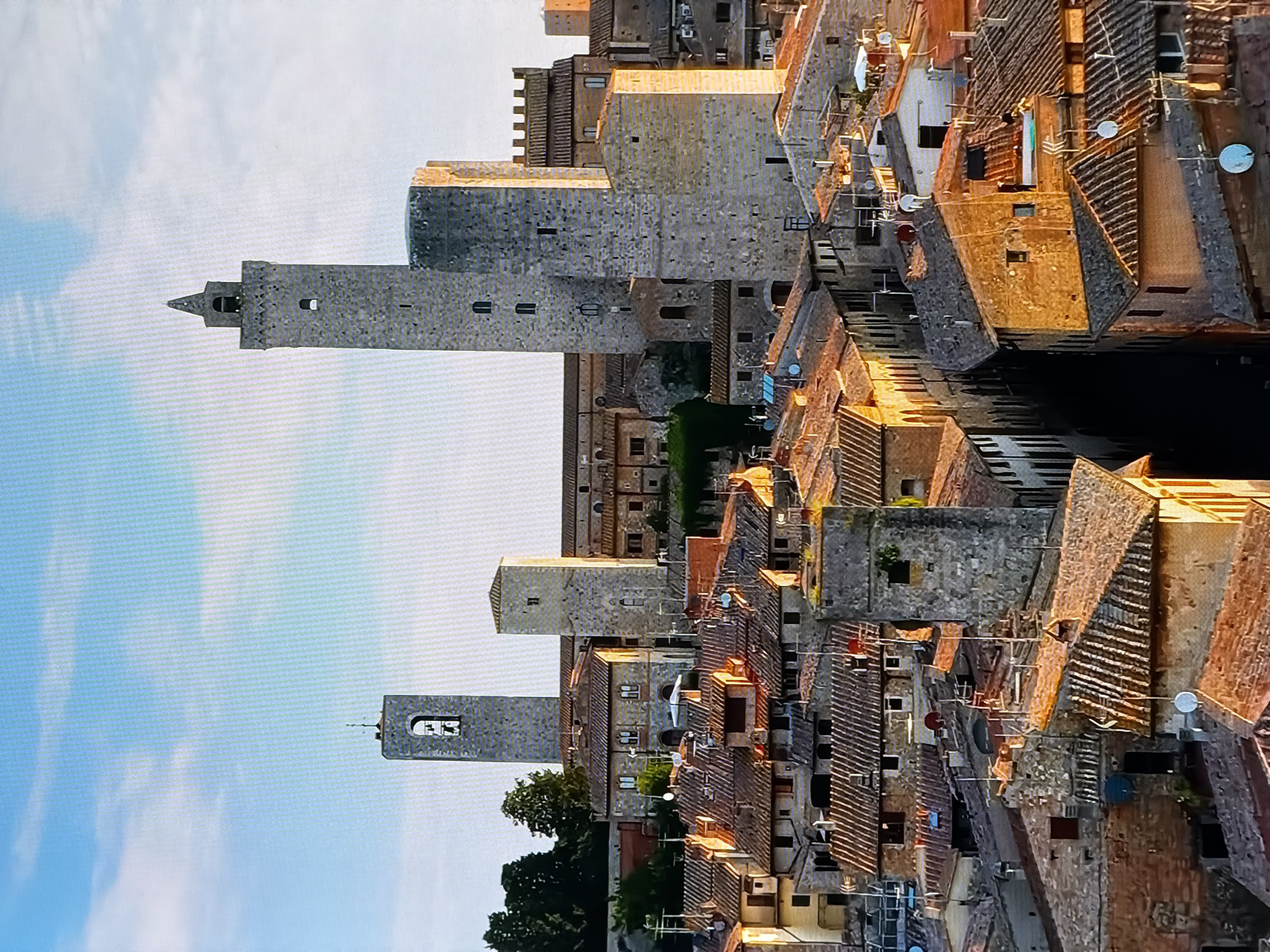 This is a daytime, aerial shot of the historic town of San Gimignano, Tuscany, Italy, not Chirchik, Uzbekistan. The scene is dominated by numerous medieval towers and buildings with terracotta roofs, densely packed together. The architecture is primarily stone, with some stuccoed facades. Sunlight casts long shadows, indicating it's likely late afternoon or early morning. The sky is a clear blue with scattered white clouds. There are no people or discernible actions visible in the image. The focus is on the urban landscape and its preserved historical character.
