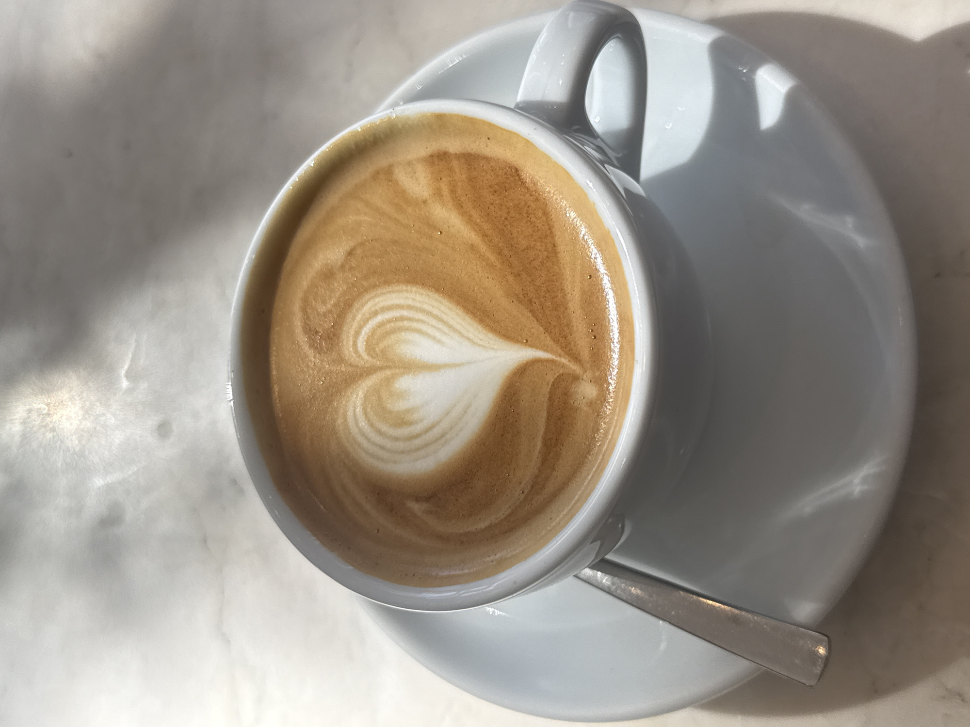 A white ceramic coffee cup, filled with a latte featuring a distinct heart-shaped latte art, is centrally positioned on a matching white ceramic saucer. A silver-colored metal spoon rests on the saucer, adjacent to the cup. The ensemble is placed on a light-toned, possibly marble, surface exhibiting subtle grey marbling and reflections from an overhead light source. Strong directional lighting from the upper left casts defined shadows of the cup and saucer onto the surface. No individuals are present, and no actions or interactions are depicted. The location is contextualized as an indoor setting within the city of Cape Town, South Africa.