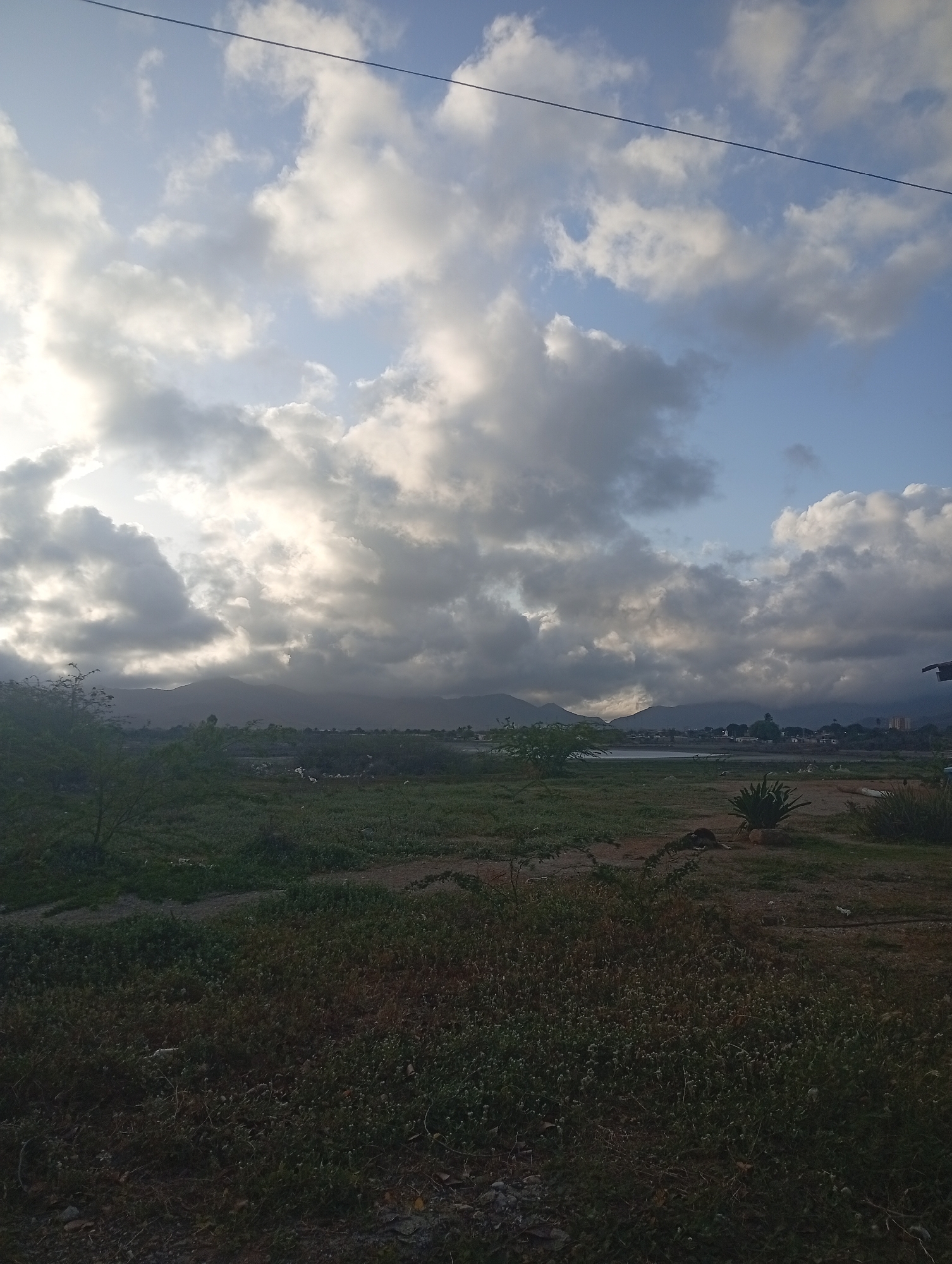 The image depicts a landscape scene in Juan Griego, Venezuela, under a sky filled with dramatic, mostly grey clouds and patches of blue. In the distance, a mountain range is faintly visible. The foreground is dominated by a grassy field interspersed with dirt patches and low vegetation. Further back, a body of water can be seen along with some structures. The overall lighting suggests it could be early morning or late afternoon. A power line stretches across the top of the image. There are no people visible in the frame.
