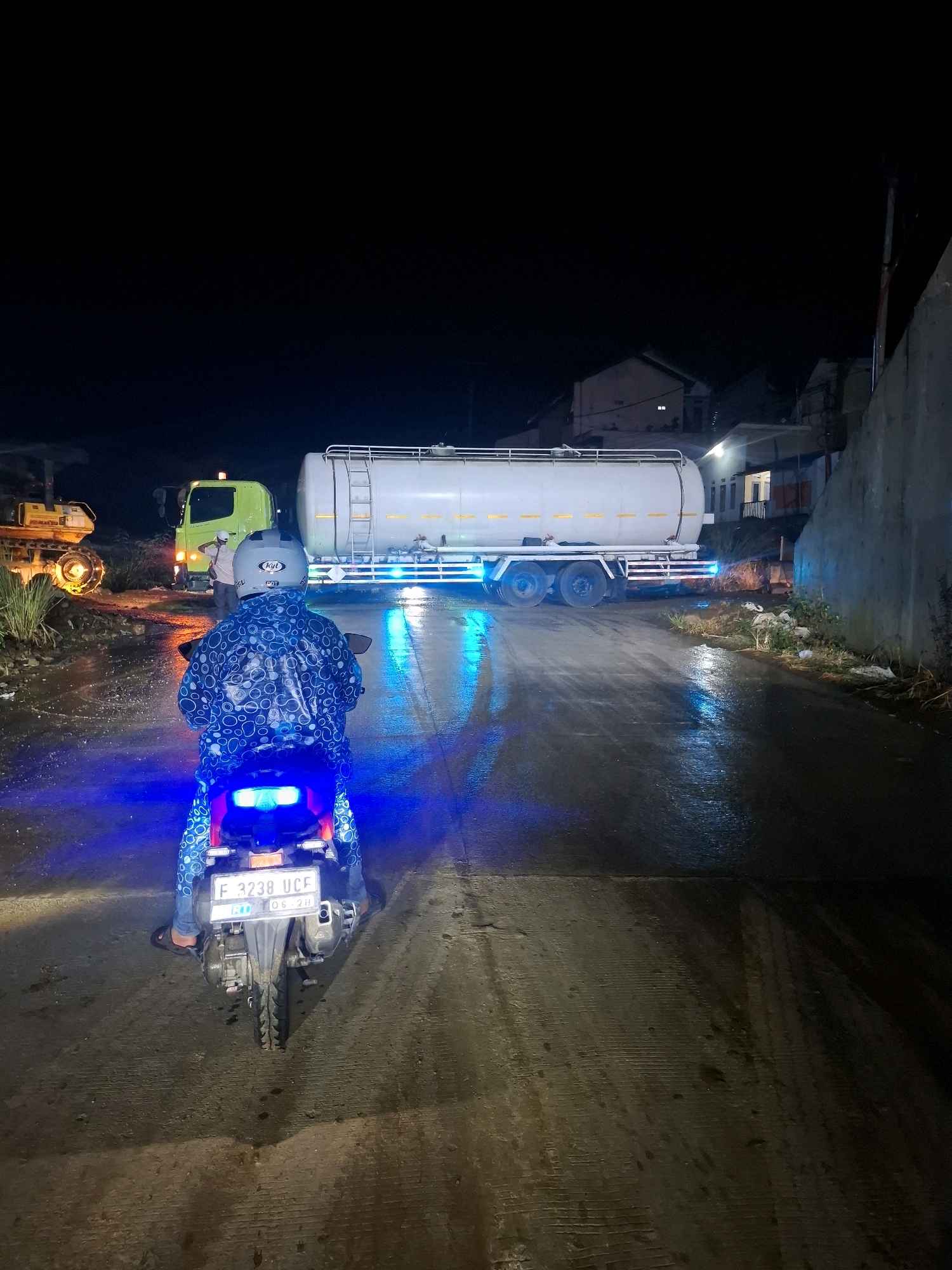 At night in the city of Ciheulang Tonggoh, Indonesia, a person on a motorcycle wearing a blue rain jacket with a white circular pattern is facing away from the camera, looking at a large white tanker truck. The tanker truck is illuminated by blue lights, which are also reflected on the wet asphalt road. To the left of the tanker truck, a yellow construction vehicle with its lights on is visible. A bright green truck is parked behind the construction vehicle, with a person standing near it. The road appears to be wet and dirty, with debris scattered along the sides. In the background, several houses with illuminated windows are visible. The overall scene suggests a busy industrial or construction area at night, possibly after rainfall.