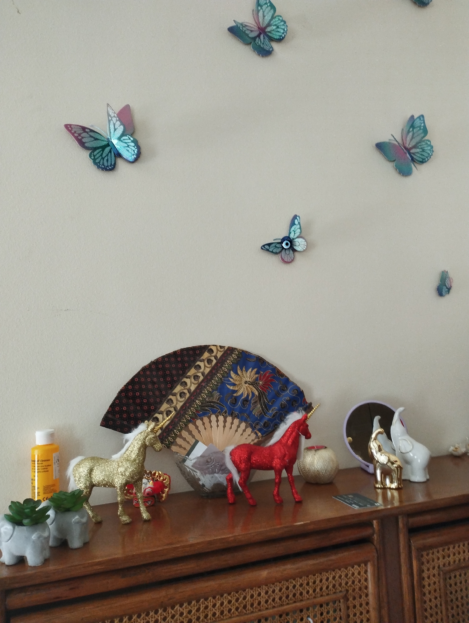 The image shows a wooden dresser with several decorative items on top. The dresser has a woven front. The objects on the dresser include two small grey elephant-shaped planters with succulents, a yellow bottle of liquid, a gold glitter unicorn figurine, a red glitter unicorn figurine, a small round candle holder, a folded fan with a patterned design, a small gold elephant figurine, and a white elephant figurine. Behind the dresser, on a plain, light-colored wall, are several decorative butterflies. These butterflies are in various shades of blue and teal, with some having pink accents. One butterfly has an eye design on its wings. A small, dark object is visible on the wall between two butterflies. The lighting suggests it is daytime, likely indoors.