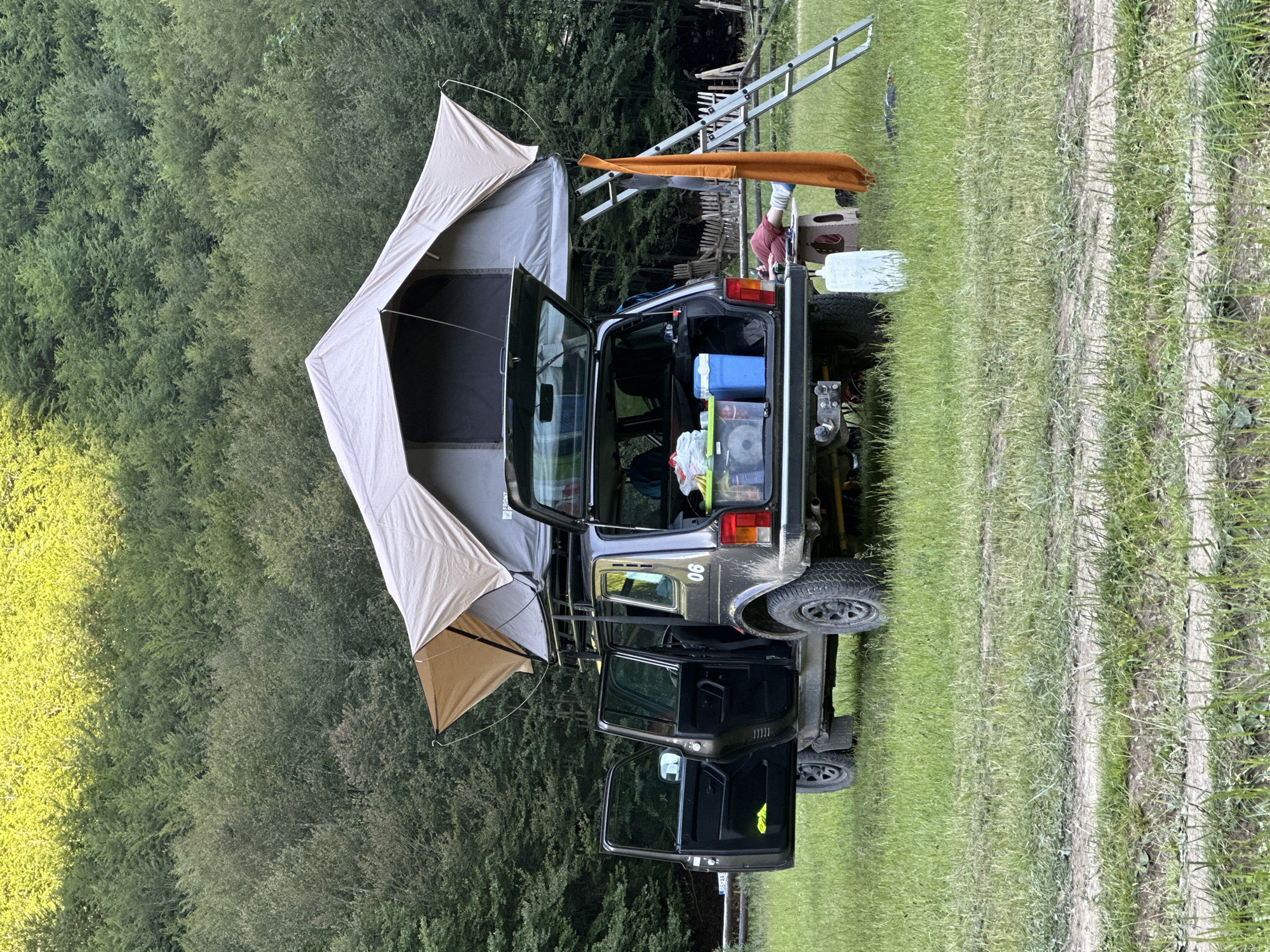 A dark gray Lada Niva vehicle is parked in a grassy field, with its rear cargo door and all four passenger doors open. A tan rooftop tent is deployed on the vehicle's roof rack. The tent is partially open, revealing a dark interior. The vehicle's cargo area contains several items, including a blue cooler, clear plastic bins, and bags. A silver ladder is propped against the rear of the vehicle, and an orange towel is draped over it. To the right of the vehicle, a person wearing red pants and white socks is lying down on a small stool, facing away from the camera. A white water jug is placed on the ground near the rear of the vehicle. In the background, a dense forest with green trees stretches across the frame, with the top canopy illuminated by sunlight. The ground is covered in green grass and dirt tracks. A weathered wooden fence is visible behind the vehicle. The scene appears to be set in a natural, outdoor environment.