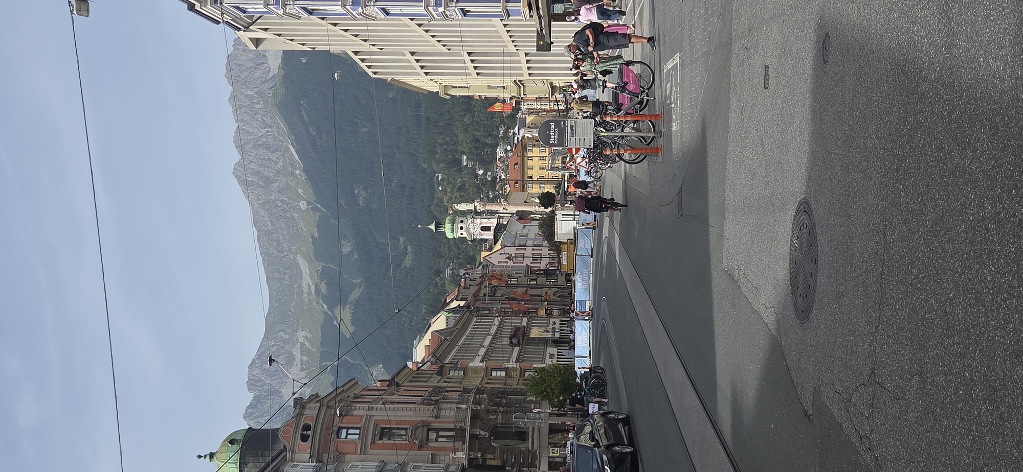 The image depicts a street scene in Innsbruck, Austria, looking towards the mountains. On the left, a multi-story building with ornate architecture and a green dome is visible. The street itself has tram tracks. On the right, a modern building with many windows and a light-colored facade stands. In the distance, a church spire with a green roof and a clock face, and a tall column topped with a statue are prominent. The background is dominated by a range of mountains covered in dark green trees and lighter, rocky peaks, under a bright, hazy sky. Several people are present, some walking on the sidewalk, others cycling. One person is riding a bicycle in the middle of the street. There are parked bicycles, including a pink one that appears to be part of a bike-sharing system, indicated by a sign that reads "StadtRAd Innsbruck". Orange cones are placed near the parked bicycles. Construction barriers are visible across the street in the mid-ground, with text that appears to say "INNSBRUCK". A black car is parked on the left side of the street, and another vehicle, possibly a taxi, is driving away from the camera. The street is paved with asphalt, and a manhole cover is visible in the foreground.