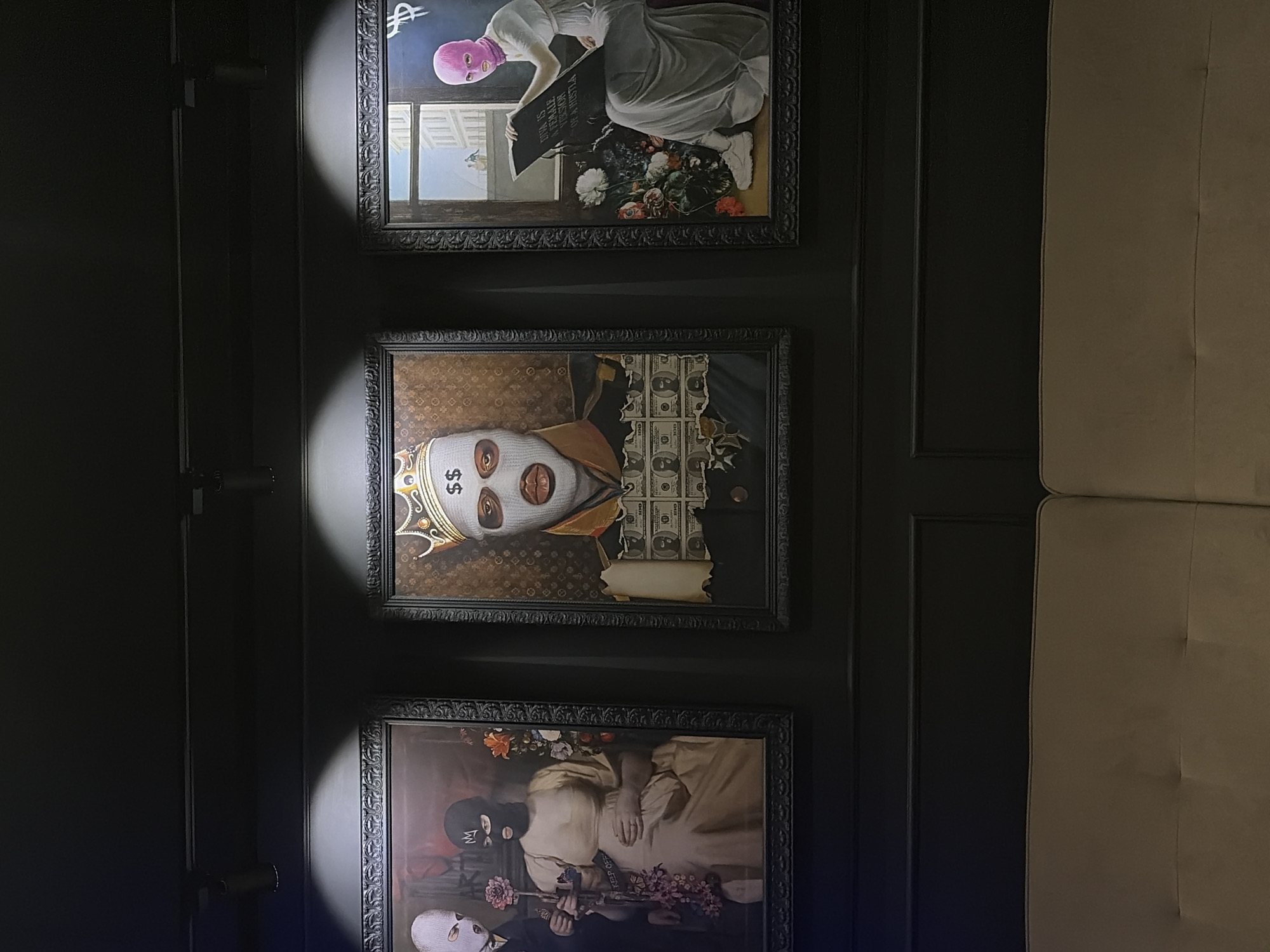 The image shows three framed artworks hanging on a dark wall. The top artwork depicts a person wearing a pink balaclava and a white dress, kneeling and seemingly writing on a gravestone. The middle artwork features a person wearing a white balaclava with dollar signs, a crown, and an expensive gold chain. The artwork has torn dollar bills behind the person. The bottom artwork depicts people in white dresses with weapons. The frames of all three artworks are dark and ornately decorated. The location is not clearly identifiable as Chaikovsky, Russia.