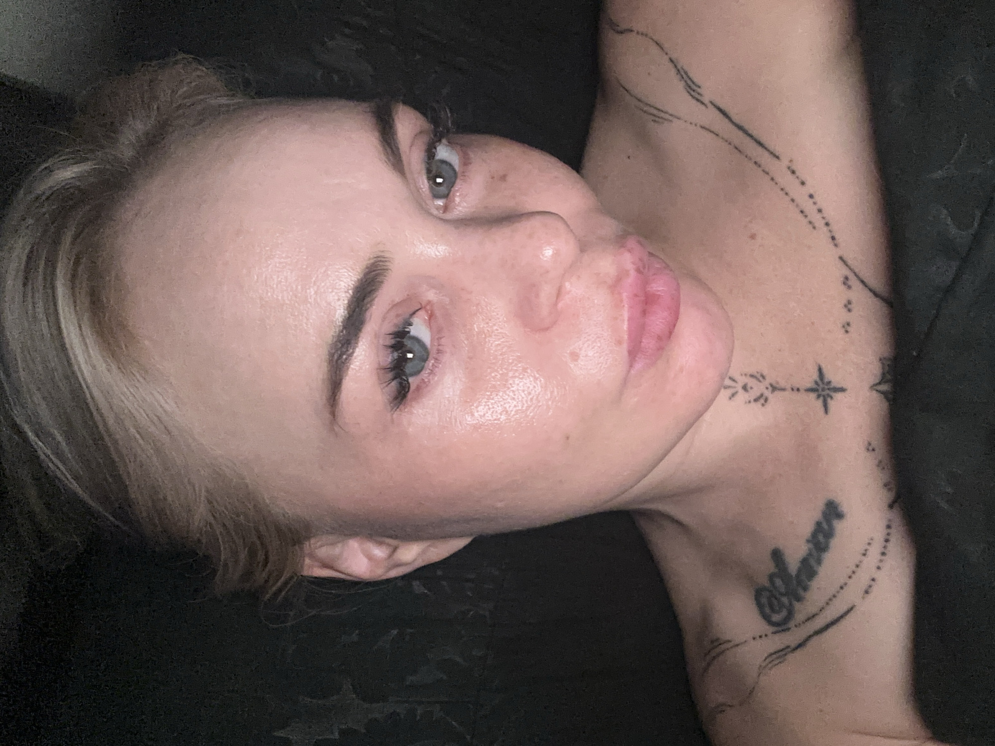 A close-up, head-and-shoulders shot shows a woman with blonde hair pulled back, wearing light makeup. Her skin appears to have a dewy, slightly oily sheen, with some freckles visible. She has light blue eyes, full lips pursed slightly, and her eyebrows are dark and well-defined. She is wearing false eyelashes. The woman has several black tattoos visible on her chest and neck. One tattoo on her chest reads "Amor". She is positioned against a dark, indistinct background, possibly a dark fabric or wall. The image is lit from the front, highlighting her facial features and skin texture. The overall impression is of a selfie taken in low-light conditions.