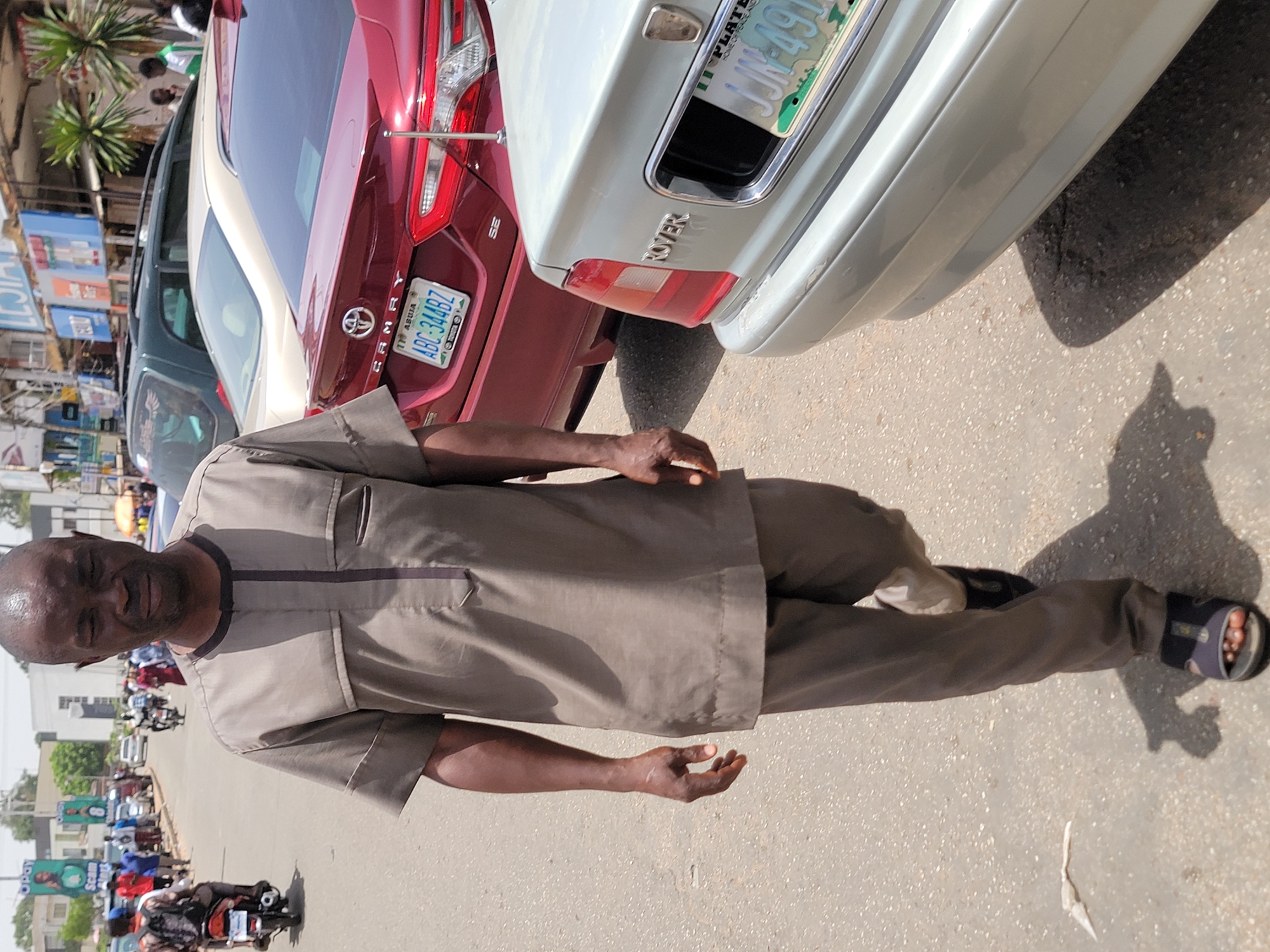 A man in a brown shirt and pants stands on a street. The background includes a red Toyota Camry with a license plate that reads "ABC-344BZ" and a white Rover with the license plate "JJN-49." The street is populated with cars and motorcycles, and there are buildings and palm trees visible in the distance. The image appears to be taken during the day. The location is Jos, Nigeria.