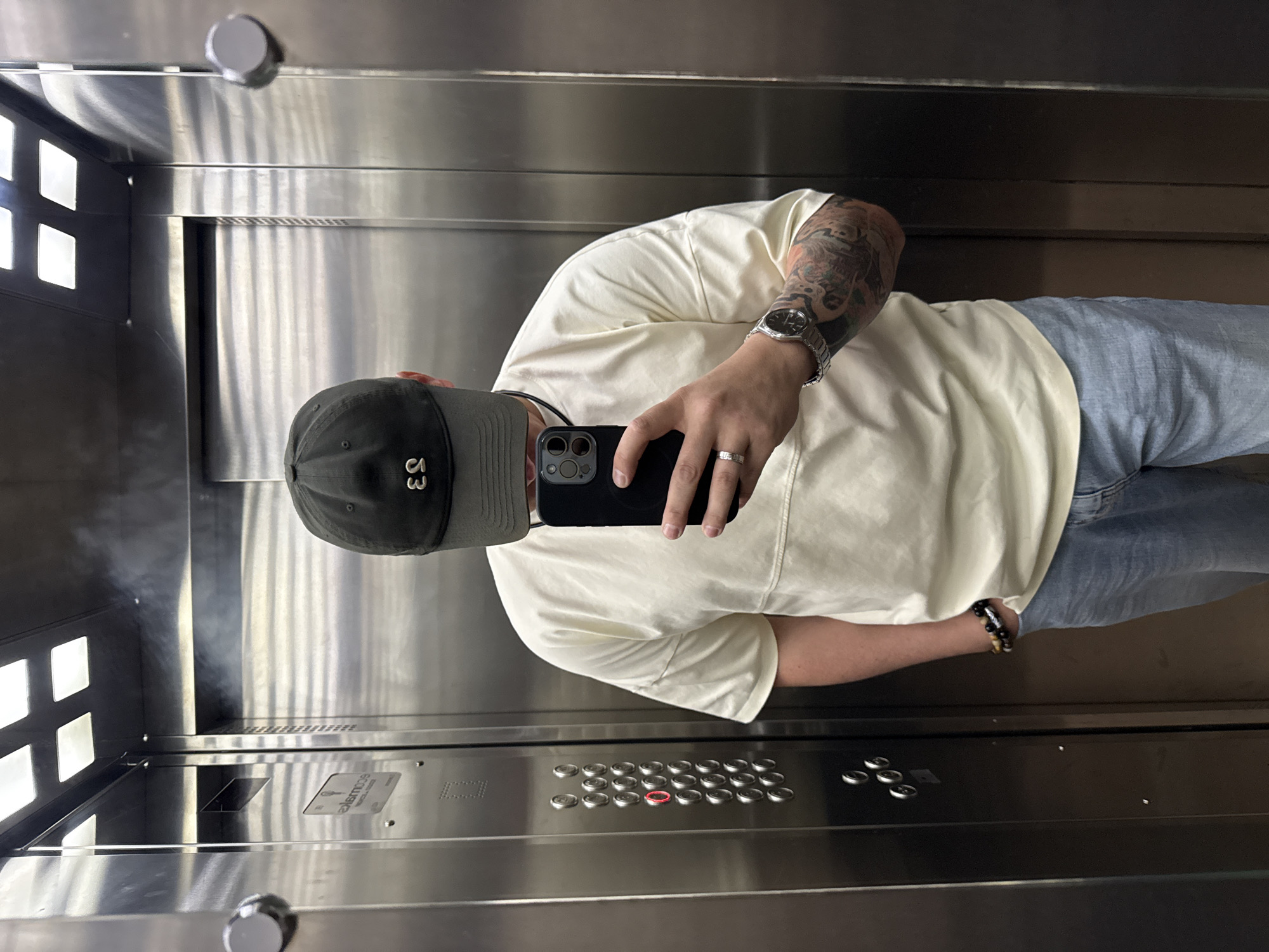 A man is taking a selfie in an elevator. He is wearing a baseball cap with the number "53" on it, a cream-colored t-shirt, and light blue jeans. He has tattoos on his left arm, a watch on his left wrist, a bracelet on his right wrist, and a ring on his left hand. He is holding a black smartphone in his right hand. The elevator has stainless steel walls and a panel of buttons. The location of the image is unknown and cannot be determined from the provided image. There is no verifiable evidence of Vladivostok, Russia in the picture.