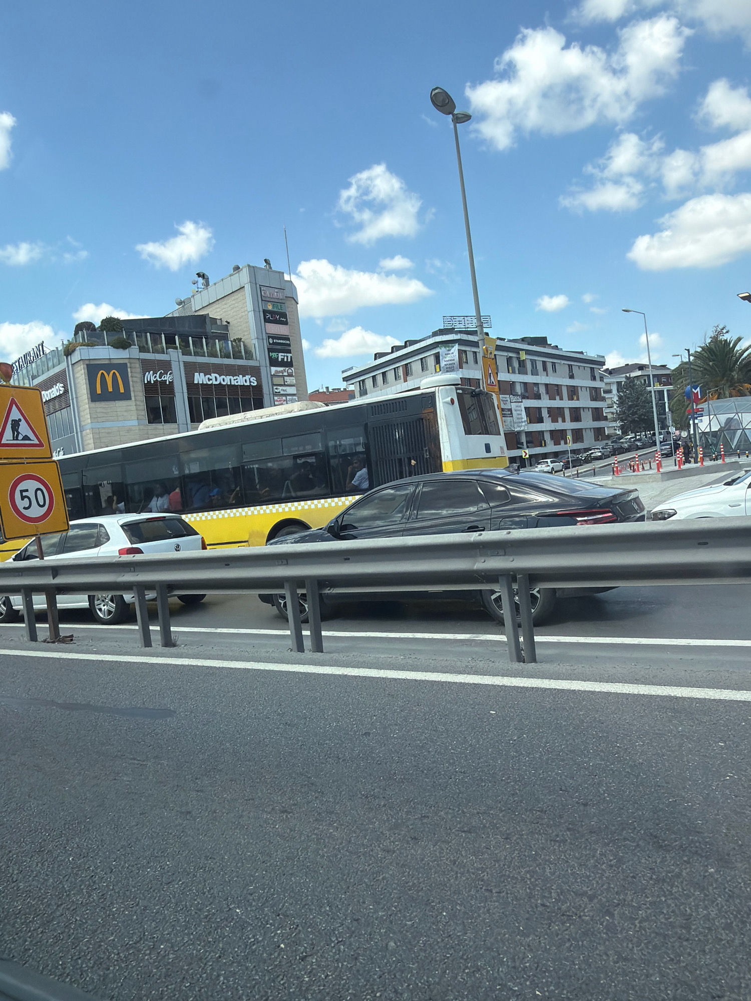 The image depicts a street scene in Bahçelievler, Turkey, likely during the daytime under a partially cloudy sky. In the foreground, a gray asphalt road surface is visible, with white lane markings. A metal guardrail runs along the road. Several vehicles are present, including a yellow and white bus with passengers visible through the windows, and a black sedan in the right lane. A white Volkswagen hatchback is seen in the left lane.
In the background, a multi-story building complex is prominent. This building appears to house several commercial establishments, including McDonald's, McCafé, and other businesses identified by signs like "FILE," "PLAY," "STARBUCKS," and "MFIT." A tall street lamp with two lights stands near the building. Traffic signs are also visible, including a sign with a worker digging and a circular sign indicating a speed limit of 50. The architecture suggests an urban environment, with palm trees visible to the right. The overall impression is one of a busy urban thoroughfare with commercial activity.