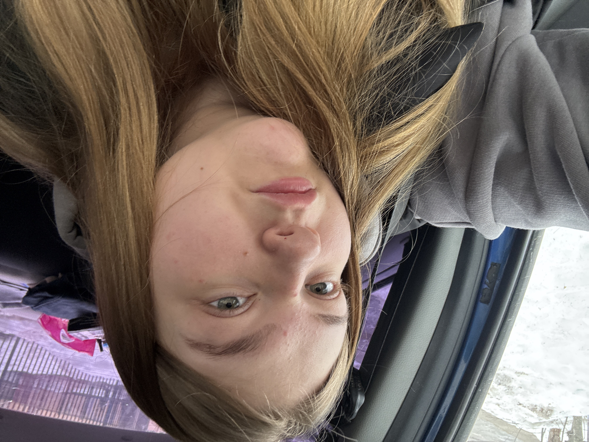 The image is an upside-down selfie of a young woman with light brown hair and fair skin. She has light-colored eyes and is wearing a gray hooded sweatshirt. The background appears to show the inside of a car, with a glimpse of an outdoor scene through the car window that appears to be wintery. Based on the context provided, the location is assumed to be Chaikovsky, Russia, given its inclusion in the prompt.