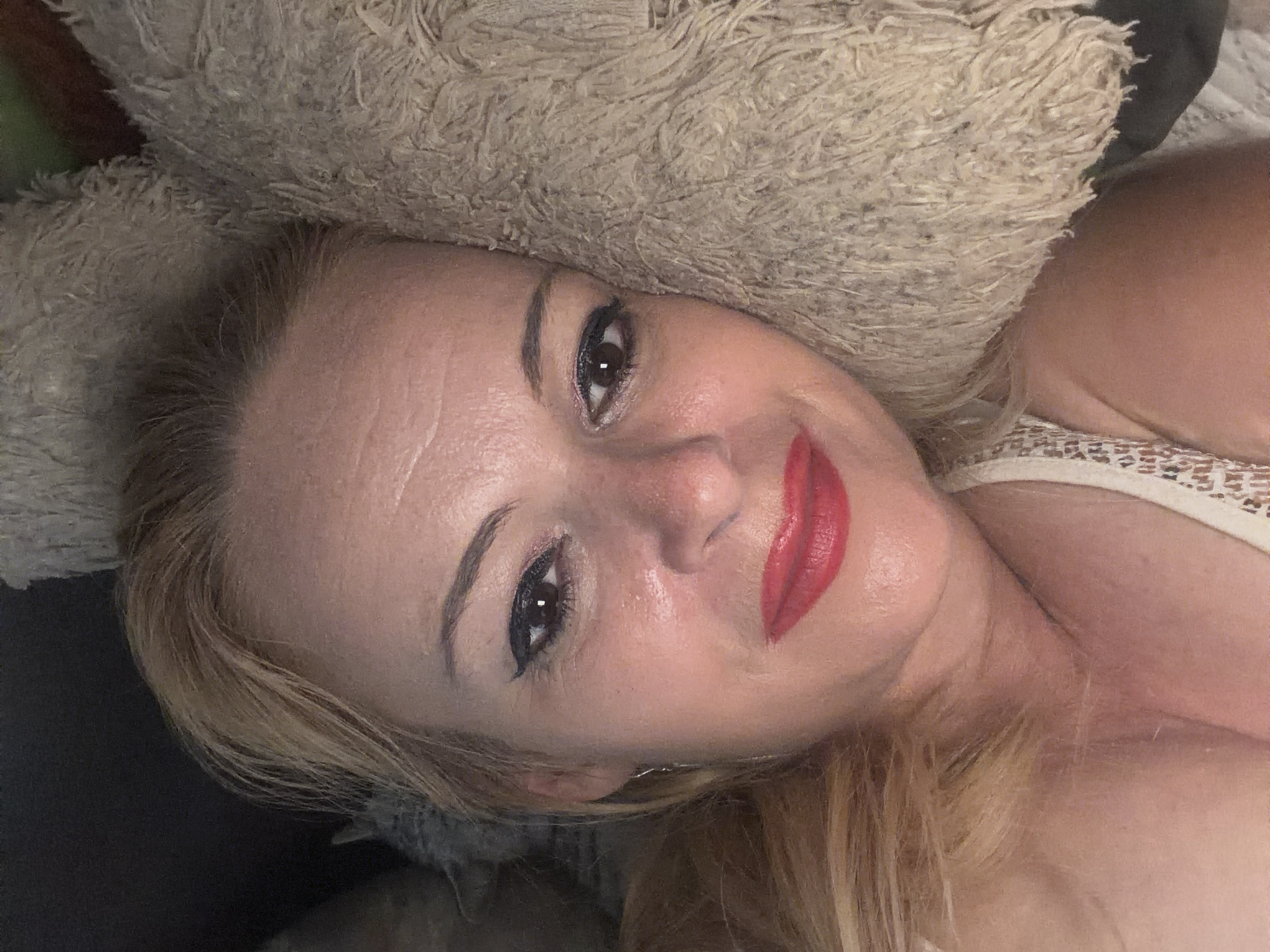 A close-up, overhead shot shows a woman with blonde hair, lying down with her head on a fluffy, beige pillow. She is wearing red lipstick and eye makeup. Her hair is pulled back slightly from her face. A dark background is visible behind her head and to the left. Her right shoulder and chest, adorned with a white lace top, are visible in the lower right of the frame.