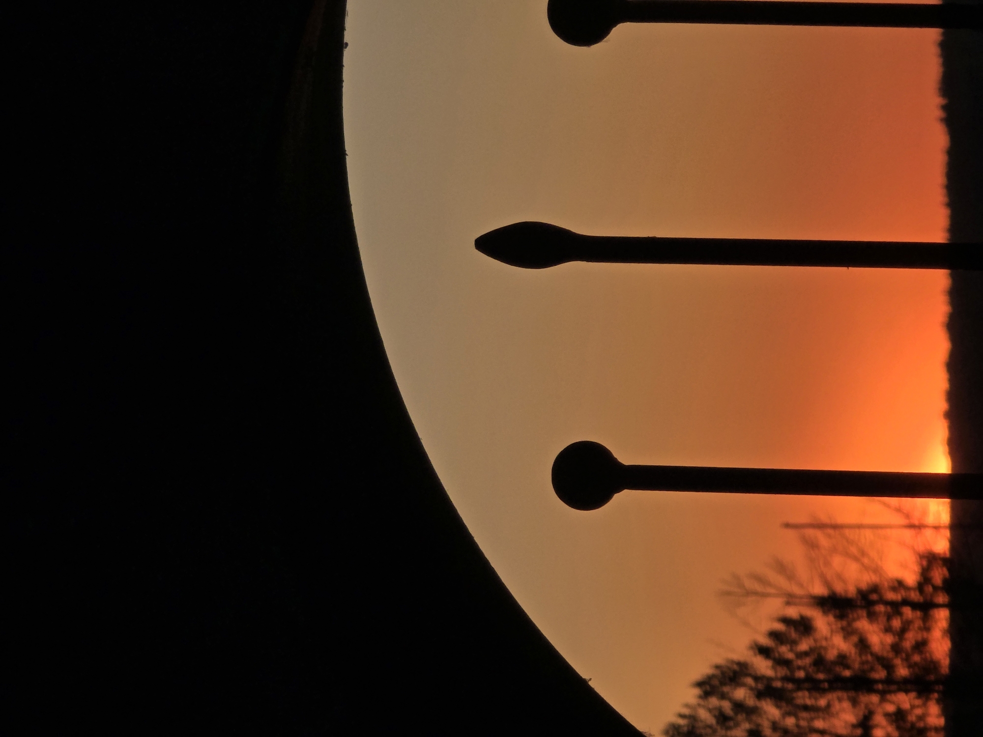 The image shows the reflection of a sunset in the city of Chaikovsky, Russia. The sunset is reflected in a curved surface, possibly a window or mirrored object. The sky is orange and yellow, with the silhouette of trees and streetlights visible. The left side of the image is mostly black.