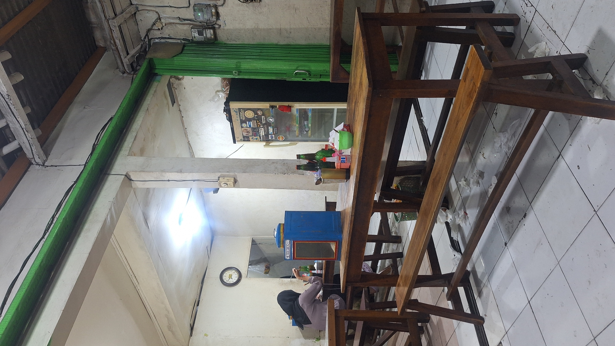 The image shows the interior of a small eatery or food stall, likely in Sukaharja, Indonesia. A woman wearing a hijab sits at a long wooden table, possibly interacting with a blue object (perhaps a dispenser or small machine) on the table. The table itself is worn and surrounded by similarly styled wooden benches. Several glass bottles and other small containers are on the table. Behind the table, there is a refrigerator with stickers. A green rolling shutter is partially visible above the fridge. The walls are white and show signs of age, with electrical wiring and a meter box visible near the ceiling. A clock is mounted on the wall. The floor is tiled in white. The lighting is a mixture of natural and artificial. Discarded paper is visible on the floor under the table.