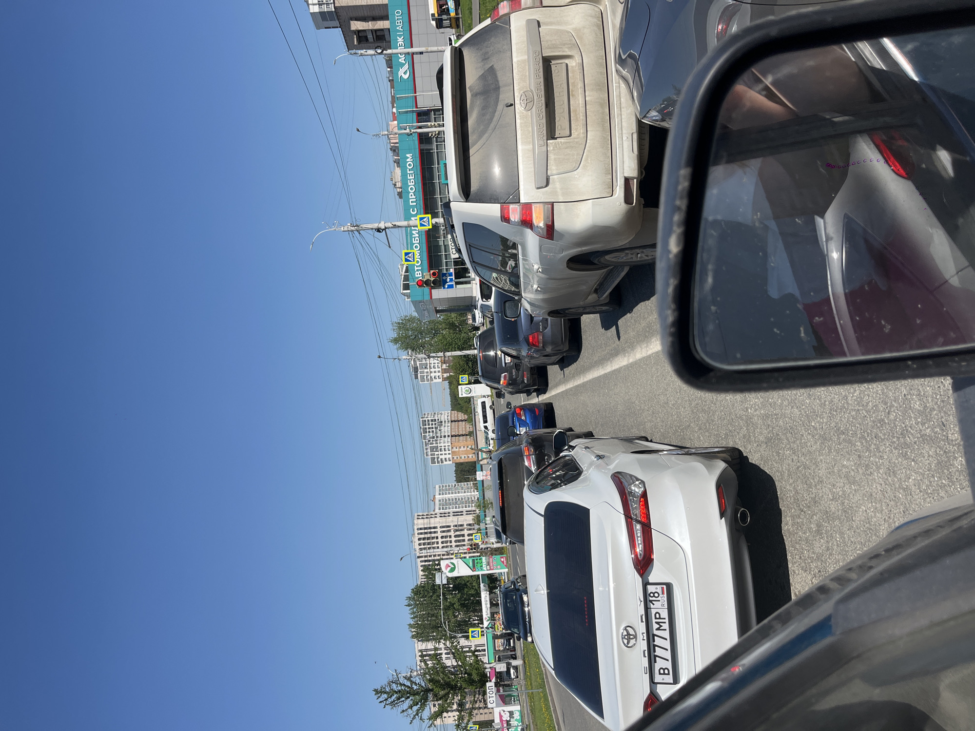 The image shows a traffic jam on a city street in Izhevsk, Russia, with several cars lined up. The vehicles are mostly passenger cars and SUVs, with brands like Toyota clearly visible. A gas station and car dealership are visible in the background with a sign that reads "LAND CRUISER PRADO" in Russian. The weather is sunny and the sky is clear blue. The vehicles are queued on a multi-lane road with city buildings in the distance. A side mirror of the car taking the photo is visible.