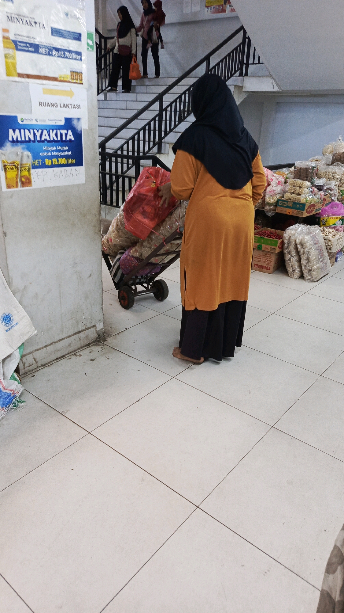 A rear view of a person wearing an orange tunic and black headscarf pushes a two-wheeled cart filled with large red and brown bags up a set of stairs. The cart has red wheels and is loaded high. The person's legs are bare from the knees down. The scene appears to be an indoor marketplace or grocery store in Ponorogo, Indonesia, based on the signage displayed on a wall to the left of the frame. The signage is in Indonesian and refers to "MINYAKITA" which is a brand of cooking oil, and "RUANG LAKTASE," meaning "lactation room." Shelves stocked with packaged food items are visible to the right of the person. The floor is tiled in a light color, and the walls are concrete or painted concrete. Two other individuals, one holding a baby, are visible further up the stairs, also heading upwards.