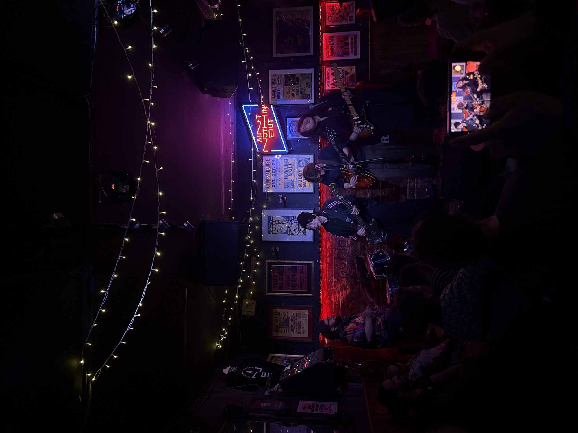 A dimly lit, slightly tilted, indoor shot captures a live music performance at a venue displaying concert posters, possibly in London, UK.
Three band members occupy a narrow stage area. The guitarist at the top wears a black long-sleeved shirt and strums a brown and black guitar, with long, dark hair. Below, a second guitarist in a dark shirt plays an orange guitar. The person at the bottom wears a dark jacket and a black beanie while playing a bass guitar. A drum set is visible to their right, illuminated by red lights.
Audience members, blurred and indistinct, are present, with one person holding up a mobile phone to record the performance. Above the performers, a neon sign reading "AIN'T NOTHIN' BUT..." is mounted on the wall, surrounded by posters advertising past concerts.
Fairy lights hang down on the left side of the frame. A sound console sits in the foreground. A person lies on the floor in front of the stage.
