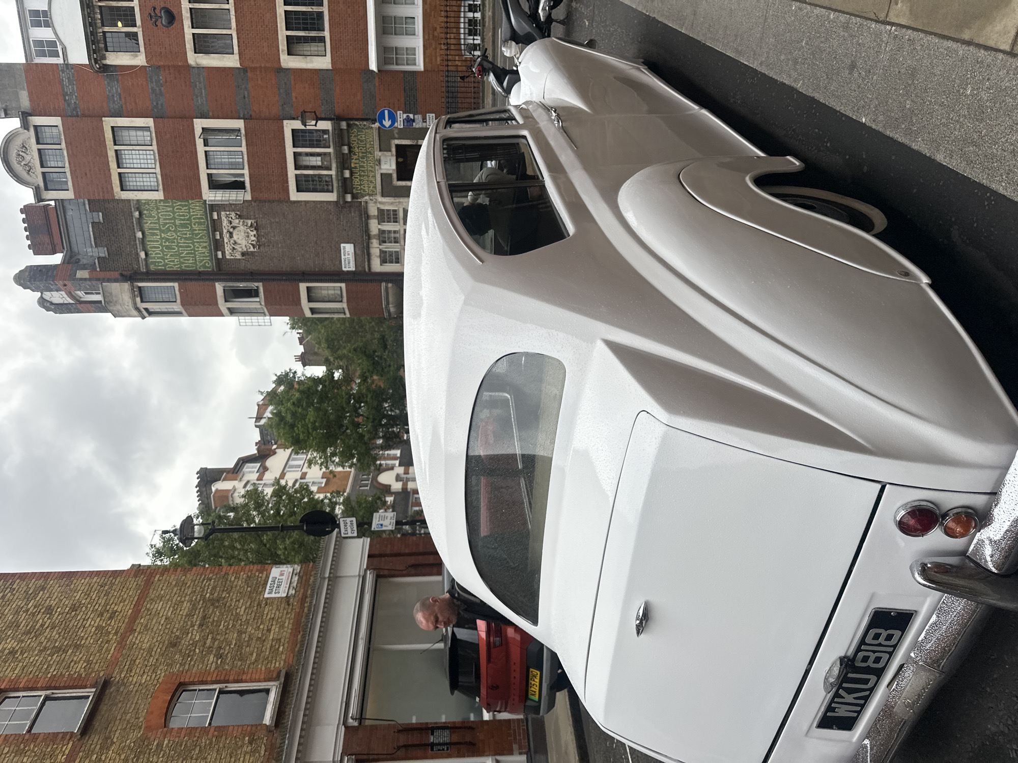 The image depicts a white classic car, possibly a vintage Rolls Royce, parked on a street in London, United Kingdom. The rear of the car is prominently displayed, with the license plate "WKU 818" visible. In the background, several buildings are seen. A sign indicates the presence of Nassau Street W1. Another sign shows "T. J. Boulting & Sons Gas & Electrical Engineers, Established 1900", The street appears to be wet, suggesting recent rainfall. In the background there is a red car with the license plate NA75 PHO.