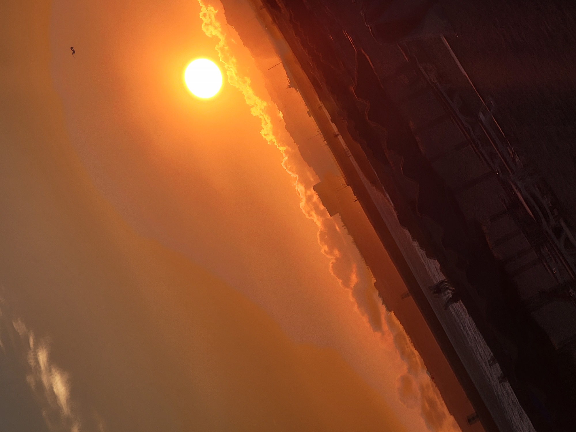 This is a vertically oriented, low-angle photograph of a sunset over the Atlantic Ocean, likely captured in Quarteira, Portugal. The sun, a bright, almost white orb, dominates the upper right quadrant of the image, casting a warm, orange glow across the sky and illuminating scattered, wispy clouds. A small, dark silhouette, possibly a bird, is visible in the upper right sky.
In the middle ground, a body of water, presumably the ocean, stretches diagonally from the lower left to the upper right. Along its edge, silhouetted structures suggest a coastline with buildings and possibly a harbor. Some faint lights are visible on the distant shore.
The foreground is occupied by a dark, silhouetted area that appears to be a beach or a boardwalk with some elevated structures, possibly awnings or beach huts, casting shadows. A few indistinct human figures are visible as dark silhouettes in the middle ground, near the water's edge, appearing to be walking or standing. The overall impression is one of a serene, end-of-day scene with a dramatic natural light display.