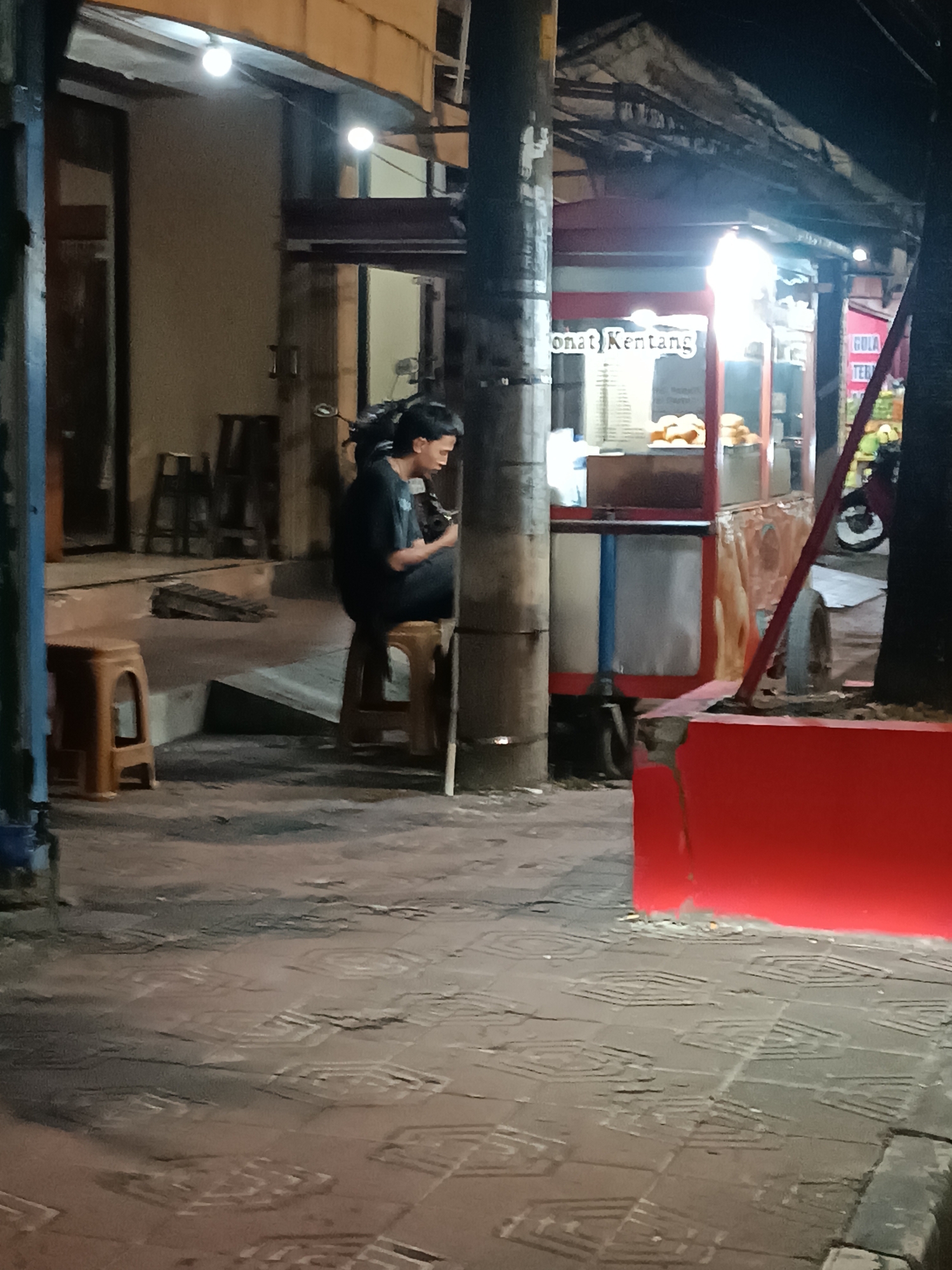 A young man with dark hair is sitting on a brown plastic stool, playing a guitar. He is wearing a black t-shirt and is facing towards the right of the frame. He is located on a tiled sidewalk, which has a repeating hexagonal pattern. To his right, a food cart with a red and white canopy is visible. The cart appears to be selling snacks, with trays of food displayed under bright lights. The text on the cart, though partially obscured, appears to read "Donat Kentang" (potato donuts). Behind the food cart, a red motorcycle is parked. The background consists of buildings with large windows and open doorways, illuminated by artificial lights. The scene is set at night.