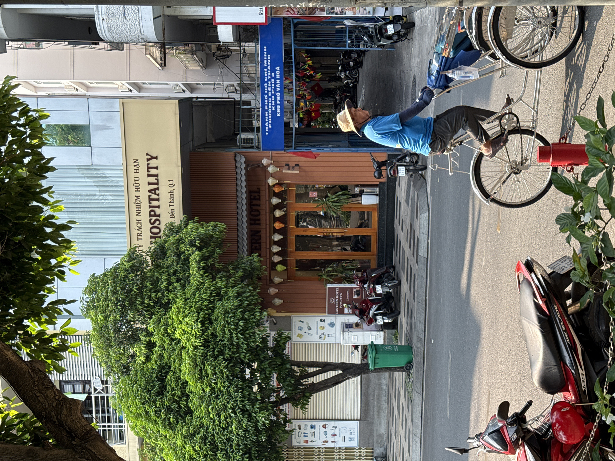 The image shows a street scene in what appears to be the city of Thủ Đức, Vietnam, during daylight. A man wearing a blue shirt and a straw hat is pedaling a cyclo (a three-wheeled passenger bicycle). There are motorcycles parked along the side of the road, near a building identified as the Lantern Hotel. Adjacent to the hotel is a building with the sign "Hospitality" and the address "Bến Thành, Q.1". A green tree stands between the street and the hotel. Above the hotel is another establishment with a blue sign. The street has a sidewalk with a patterned tile design.