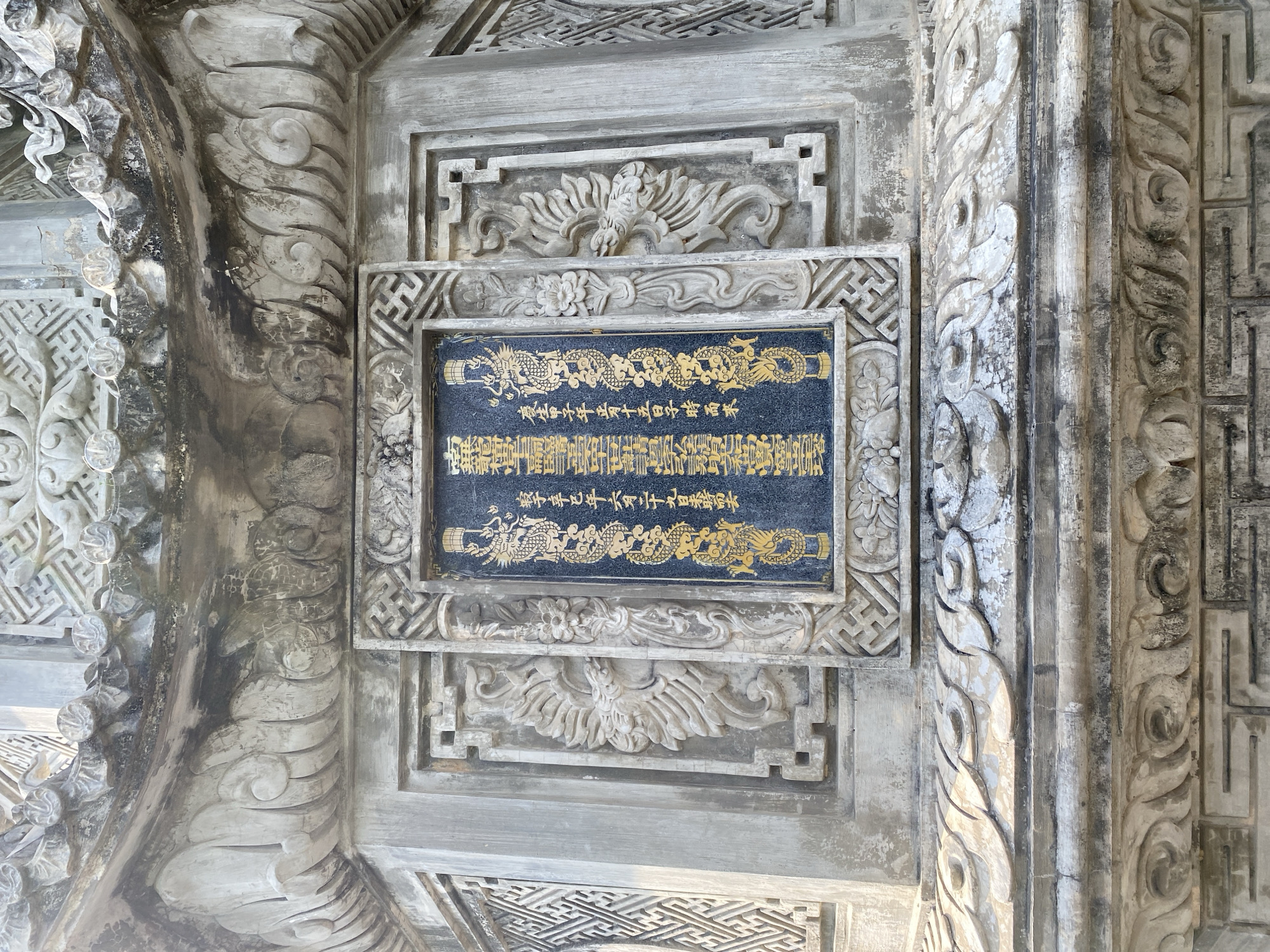 The image shows a close-up of an ornate stone carving on a structure, likely a temple or monument, located in Tay Nha Trang Ward, Vietnam. The central focus is a dark rectangular stone tablet with golden inscriptions and decorative dragon motifs. The tablet is set within an elaborately carved stone frame that features geometric patterns and stylized floral elements. Surrounding the central tablet are further stone carvings, including depictions of mythical creatures that resemble phoenixes or dragons, as well as intricate architectural ornamentation. The stone appears weathered, with signs of age and possibly some moss or dirt accumulation. The overall impression is one of historical and cultural significance, showcasing detailed craftsmanship.