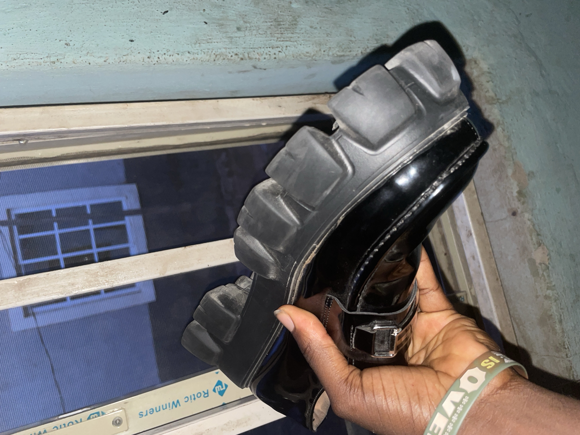 A pair of black patent leather loafers with thick, lugged soles is held in a person's hand in Jalingo, Nigeria. The shoes appear to be new. The background consists of a blue-tinted window with white panes, a beige window frame, and a textured teal wall. A green and white bracelet is visible on the person's wrist. The image is taken in an indoor setting.