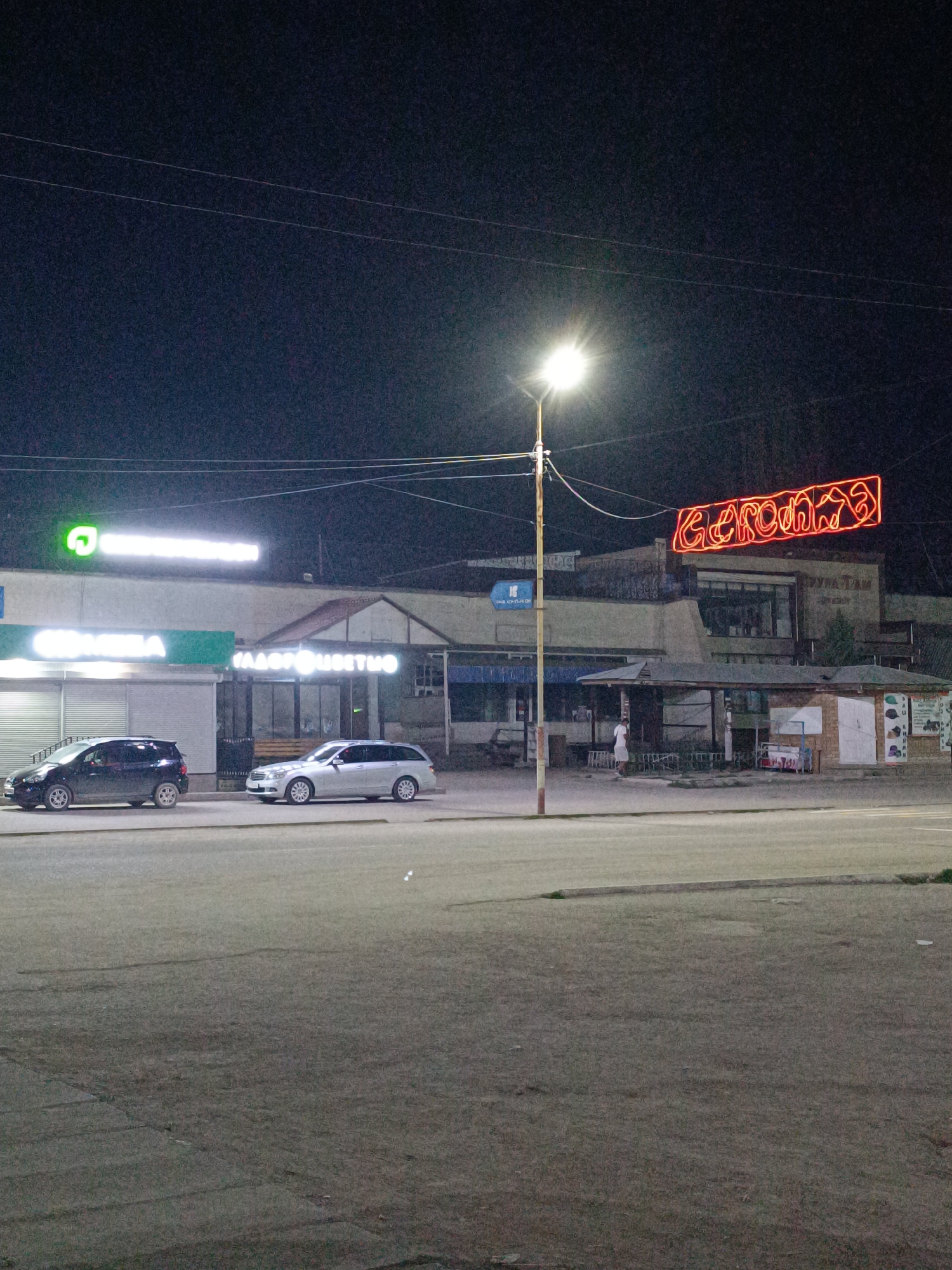 The image depicts a street scene at night in Bökönbaev, Kyrgyzstan. A brightly lit neon sign in red, reading "BEKOНБAEB," hangs above a building. To the left, a business with a green logo and white text that appears to read "ОРТОК" is visible. Further to the right, another business displays white text that reads "САДОВО ЦВЕТИНО". Two cars are parked on the street: a dark hatchback on the left and a silver station wagon on the right. A street lamp illuminates the area, casting a bright light. A single person is standing near a small kiosk or structure on the right side of the scene. The environment appears to be urban or semi-urban, with buildings and pavement. The sky is dark, indicating nighttime.
