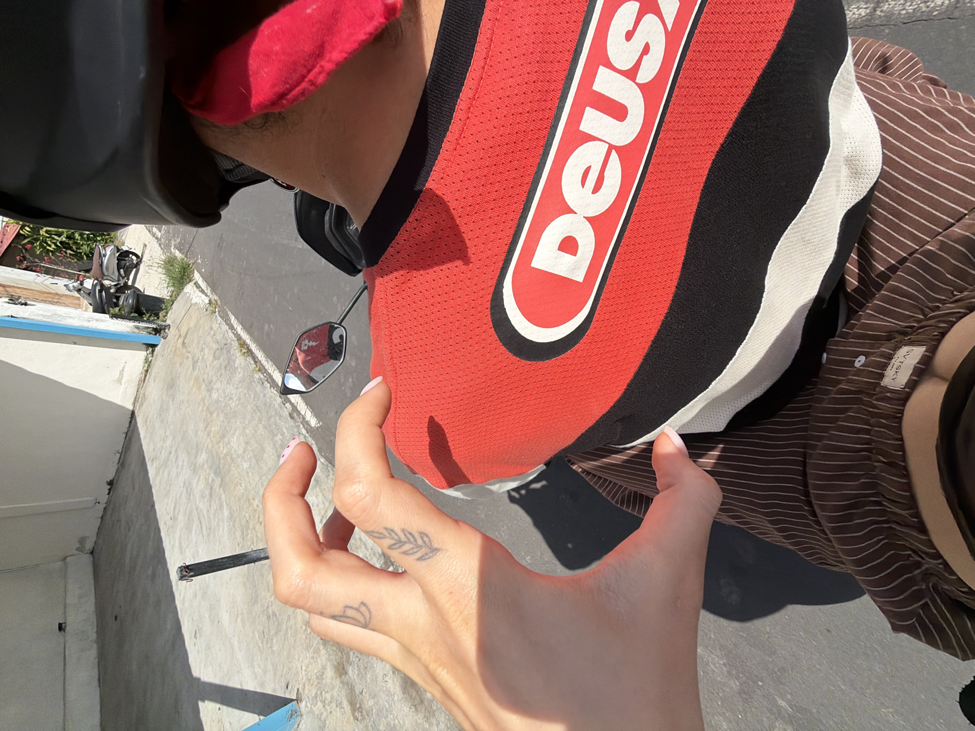 The image shows a person on a motorbike, wearing a Deus-branded top, a brown striped shirt, and a red cap under a helmet. The person's left hand is visible, showing two small tattoos. The motorbike has a rearview mirror. The scene is in North Kuta, Indonesia, as evidenced by the location data. The background includes a white building, a gray concrete surface, and a partially visible scooter. The person seems to be holding the motorbike's handlebar.
