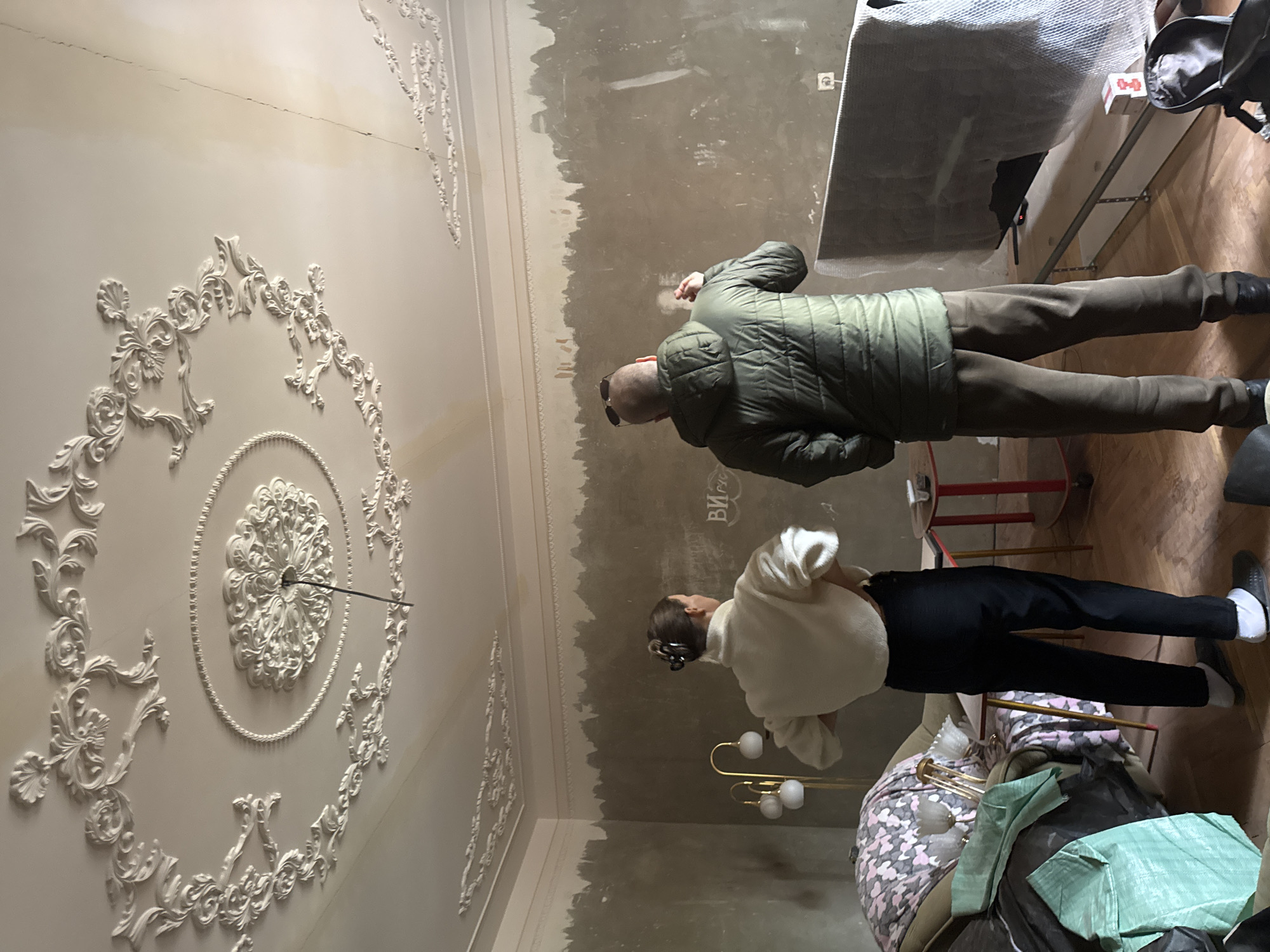 The image shows the interior of a room with ornate ceiling decorations. Two people are present: one wearing a green jacket and the other a white top and black pants. The room appears to be undergoing renovation, with unfinished walls. A lamp is visible in the scene, as well as furniture and various objects covered in what appears to be packaging material. The exact location in Chaikovsky, Russia, cannot be determined from the image alone.
