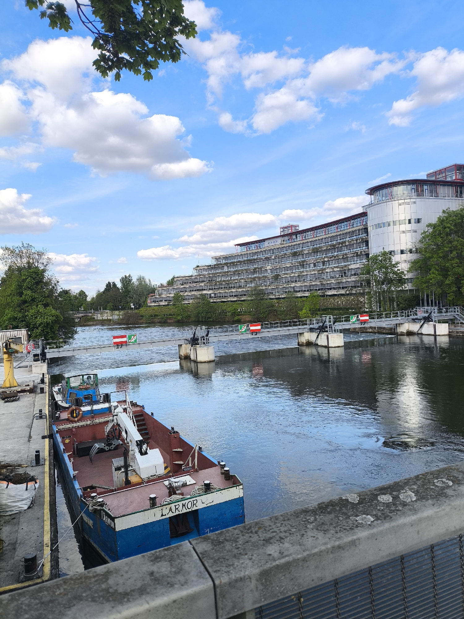 The image shows a canal or river scene in Strasbourg, France. A blue and white barge, named "L'ARMOR", is docked along a concrete quay. The barge has a crane mounted on it. In the background, a large, multi-story building with a curved roof is visible. A footbridge spans the canal, featuring red and white signs. The sky is blue with scattered clouds, and trees are present on the riverbank. A concrete barrier and metal grating are in the foreground.